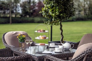 A wicker table with a tiered dessert stand, white cups, a teapot, and a vase with yellow flowers. - Woodlands Park Hotel (Cobham)