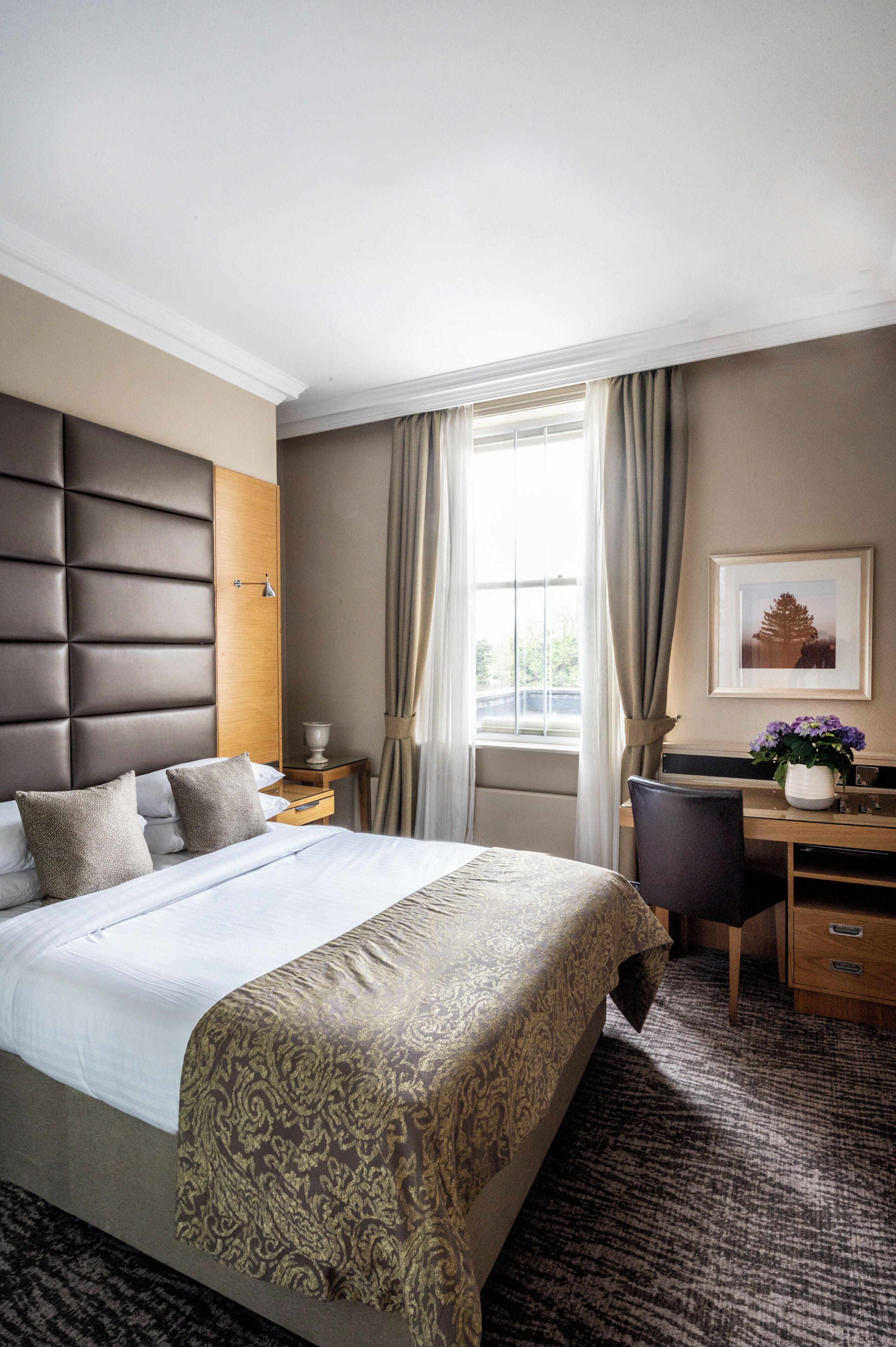 Ein Hotelzimmer mit einem großen Bett, einem Schreibtisch, einem Stuhl, einem fenster mit Vorhängen und einem Bild in einem Rahmen an der Wand.