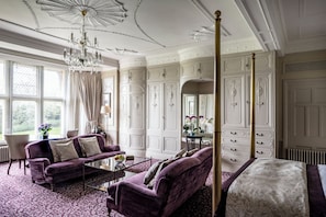 Ein luxuriöses Zimmer mit einem großen Bett, einer Sitzecke mit Sofa und Sesseln, einer Chandelüster und einem großen Fenster mit Vorhängen.