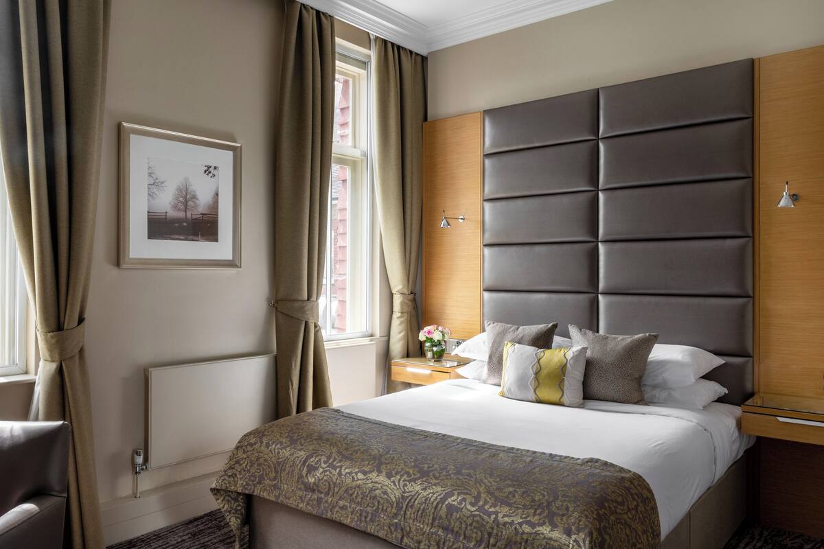 A hotel room with a large bed, a headboard, a bedside table with a lamp, a framed picture on the wall, and a window with curtains.
