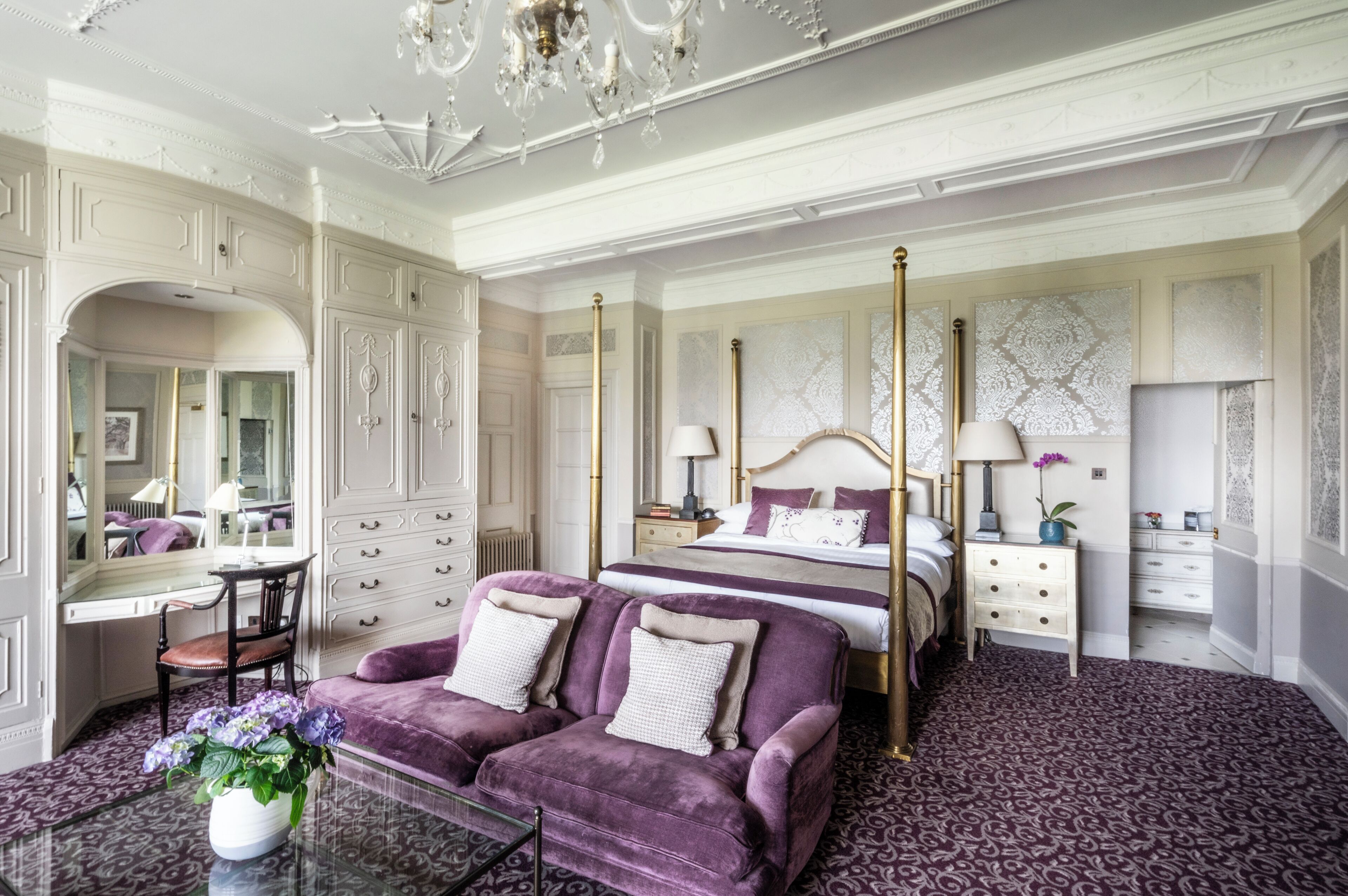 A luxurious bedroom with a four-poster bed, a vanity with a mirror, a purple sofa, and a chandelier.