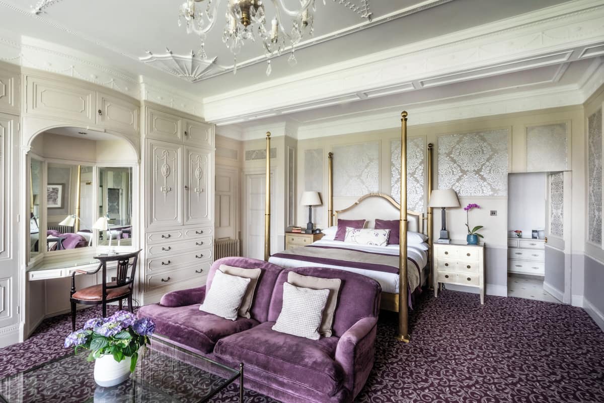 A luxurious bedroom with a four-poster bed, a vanity with a mirror, a purple sofa, and a chandelier.