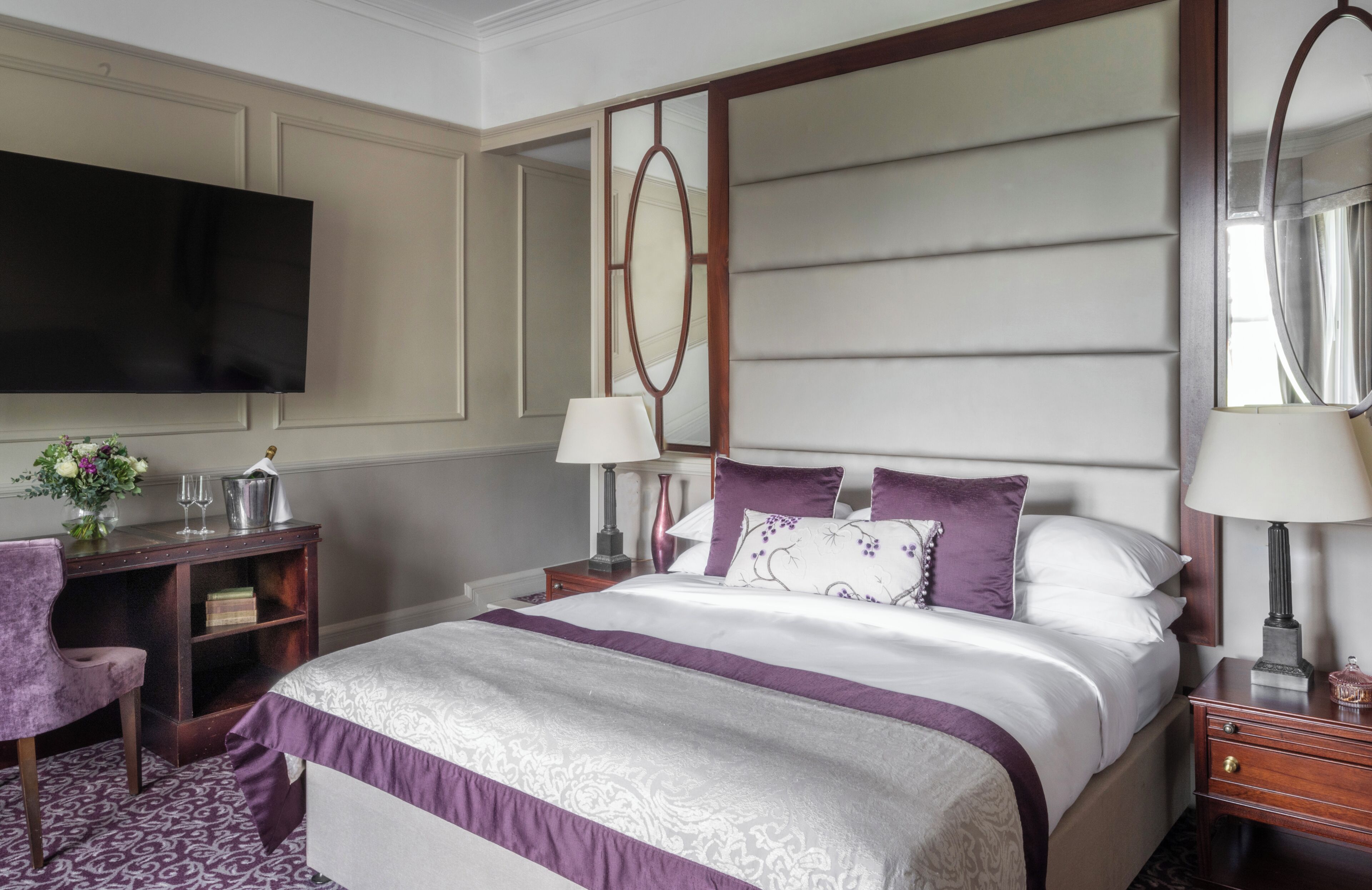 A hotel room with a large bed, a flat-screen TV, a desk with a lamp, and a chair.