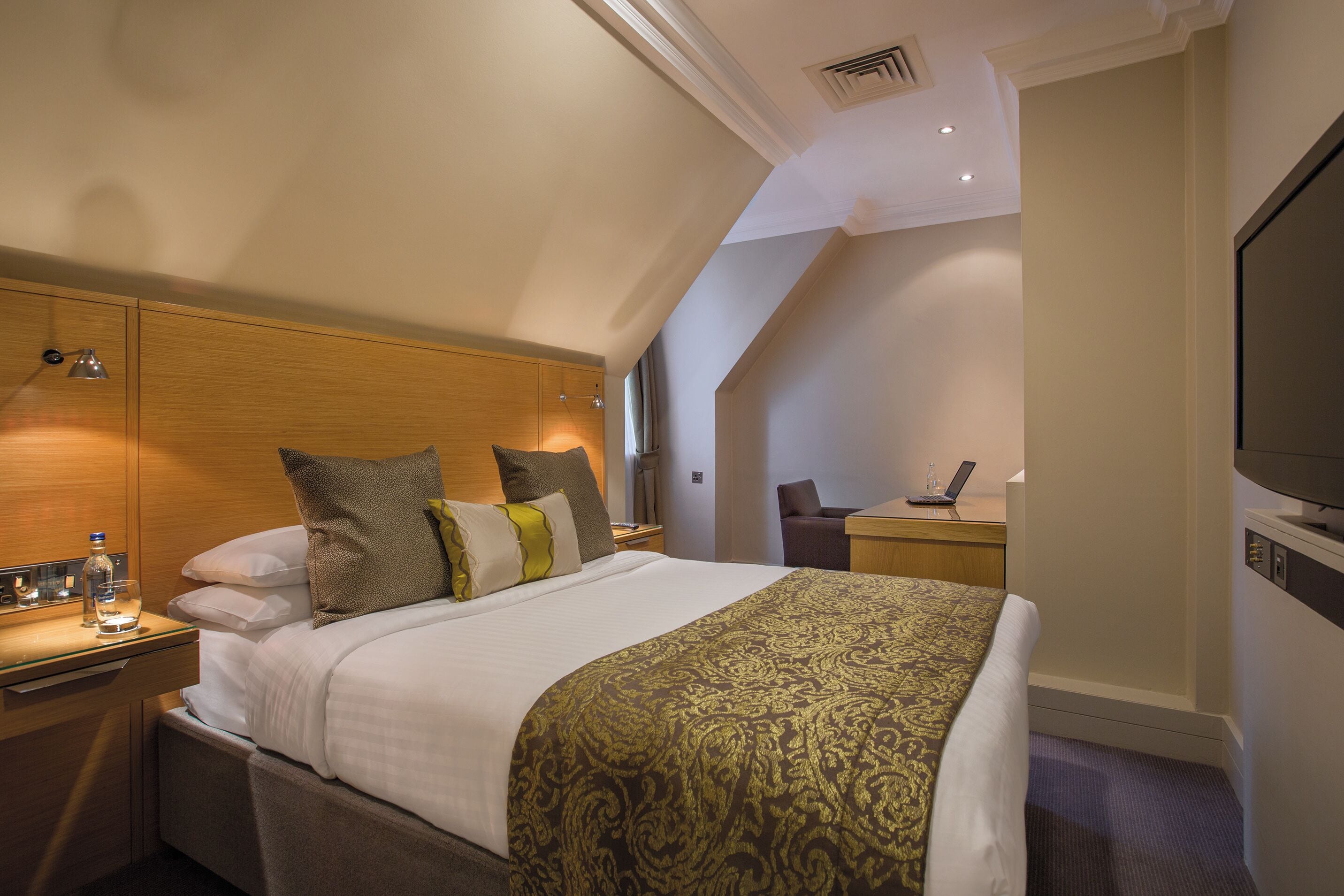 A hotel room with a bed, bedside table, lamp, TV, and a desk with a chair.