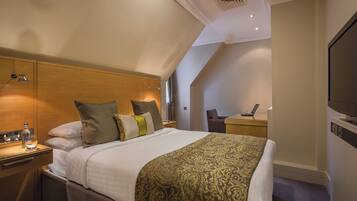 A hotel room with a bed, bedside table, lamp, TV, and a desk with a chair.