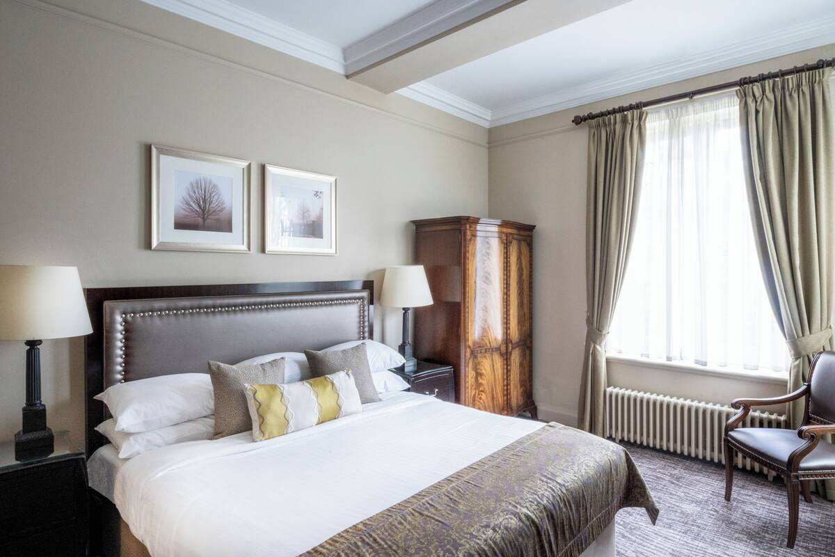 A neatly made bed with pillows, a nightstand with a lamp, a wooden cabinet, a chair, and a window with curtains.