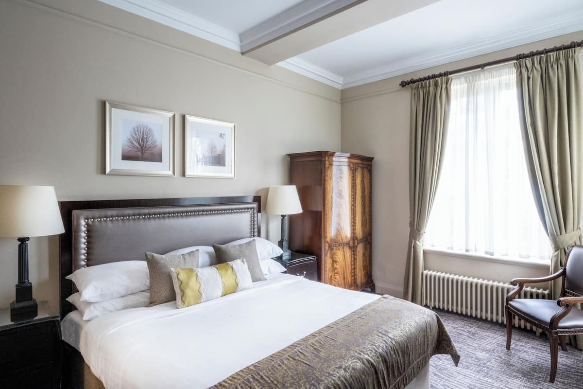 A neatly made bed with pillows, a nightstand with a lamp, a wooden cabinet, a chair, and a window with curtains.