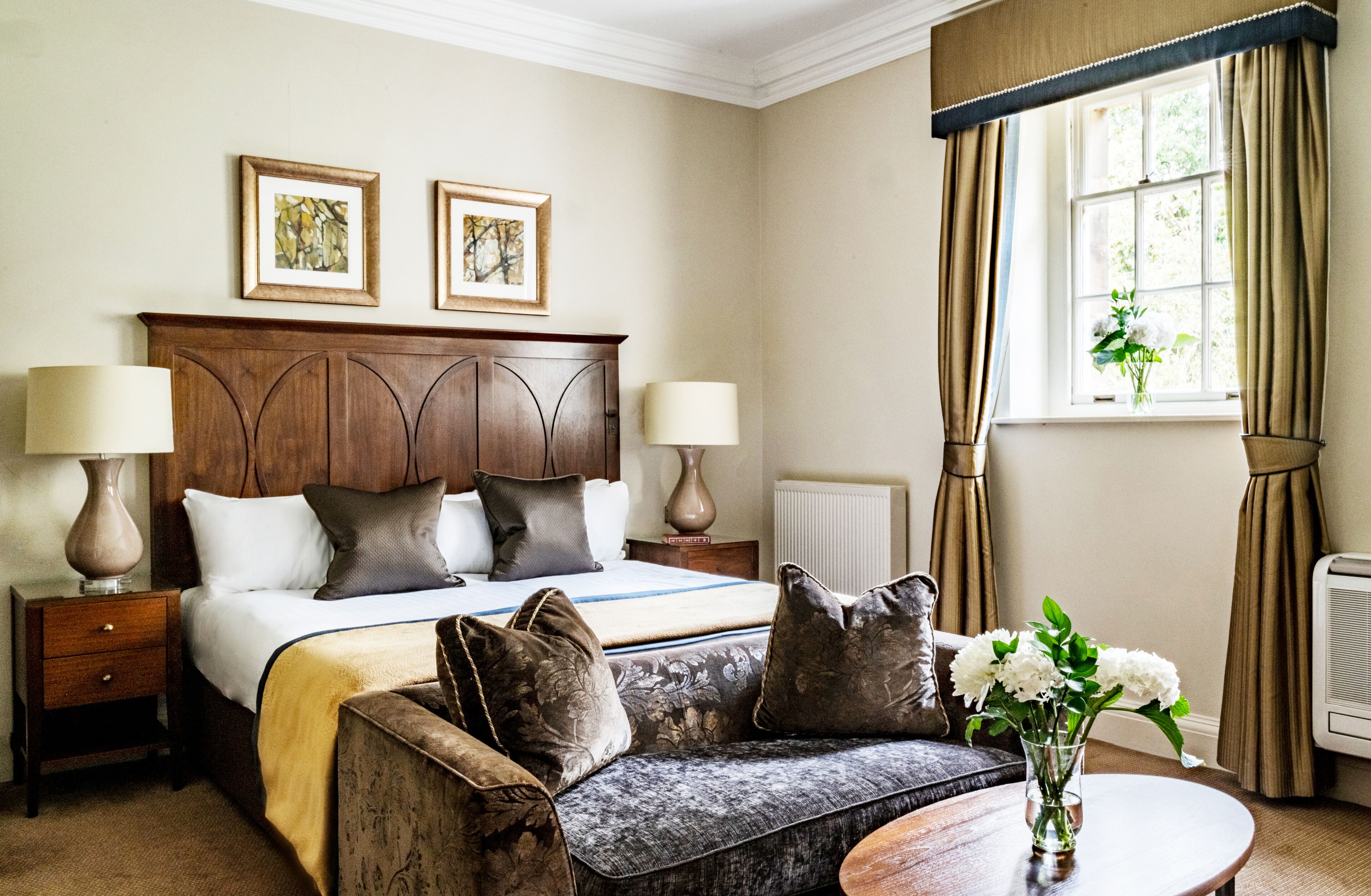 A hotel room with a bed, two bedside lamps, a sofa, a table with a vase of flowers, and a window with curtains.
