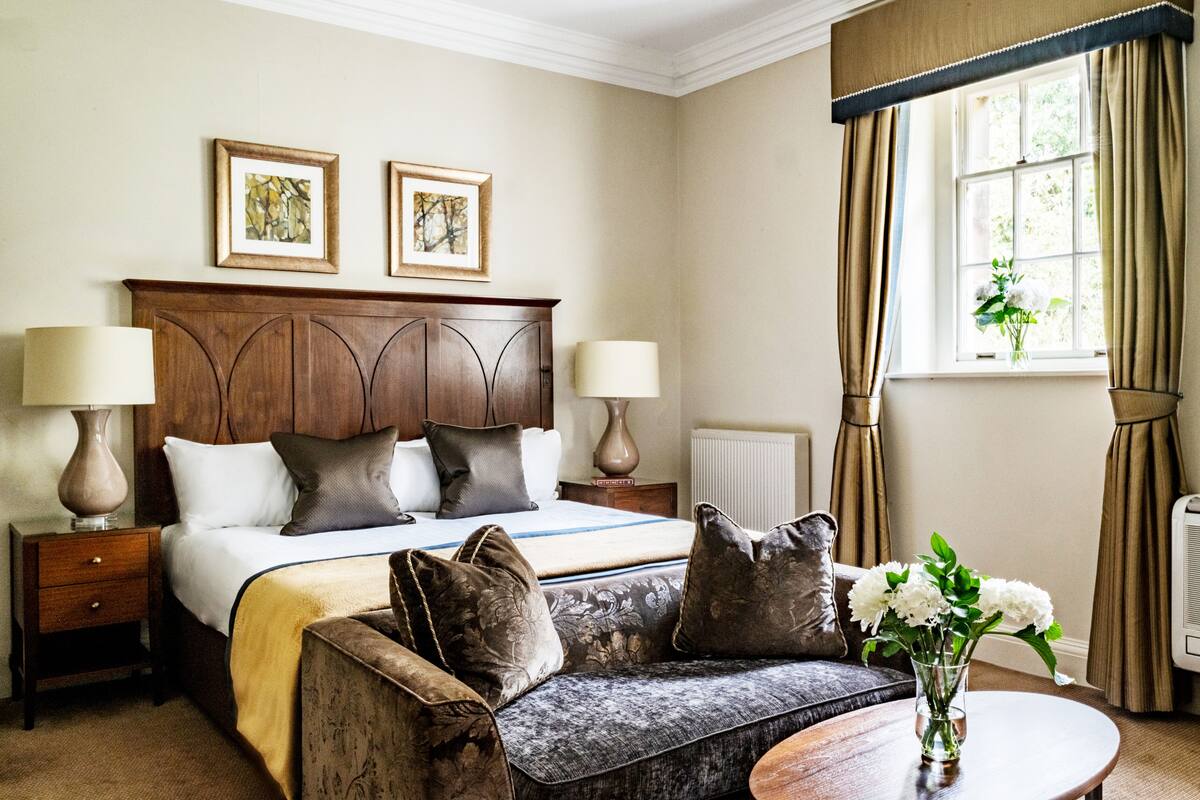 Quarto de hotel com uma cama, duas luminárias de cabeceira, um sofá, uma mesa com um vaso de flores e uma janela com cortinas.