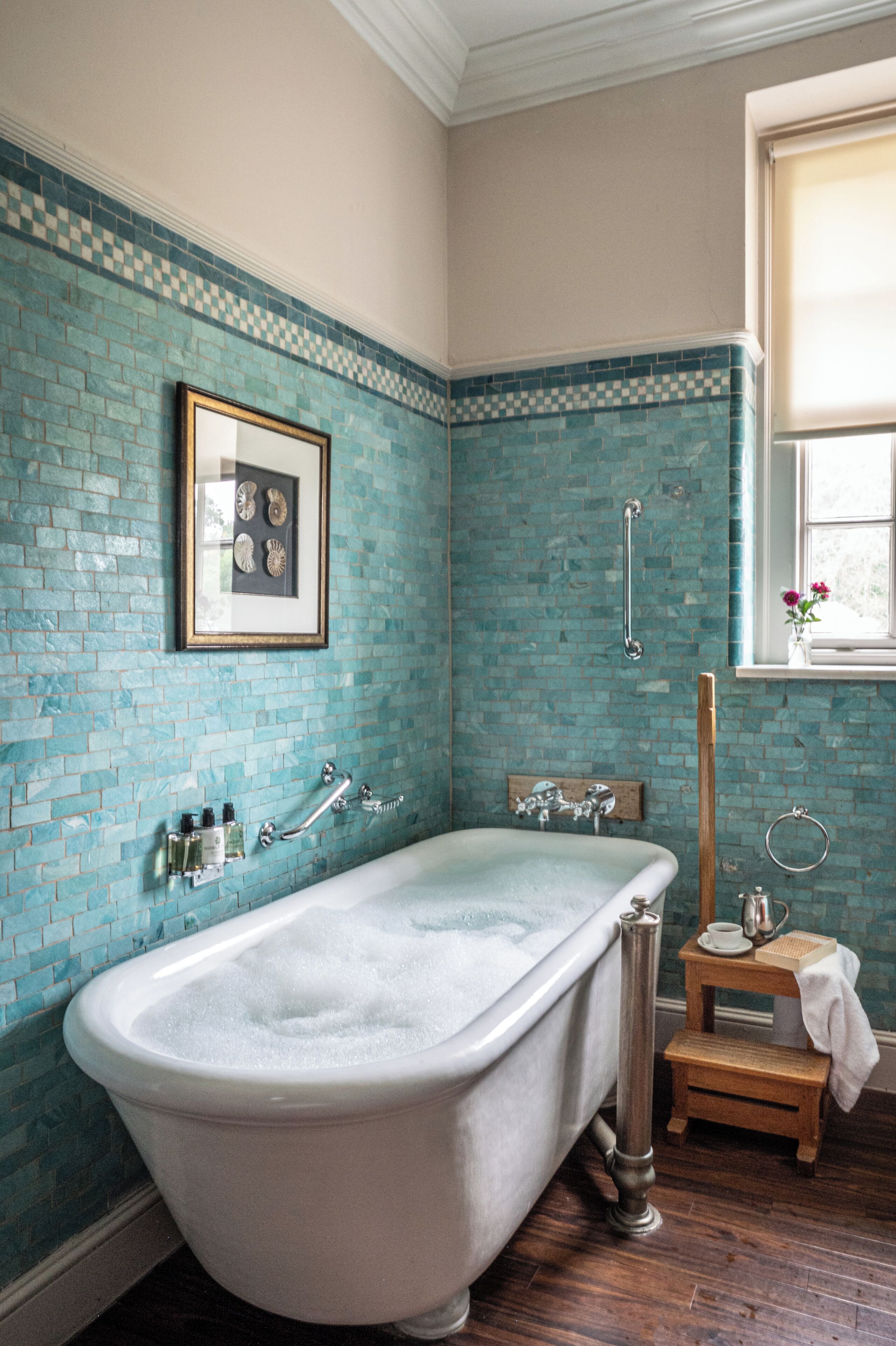 Une salle de bain avec une baignoire sur pieds, un tableau encadré au mur et une fenêtre ornée de fleurs.