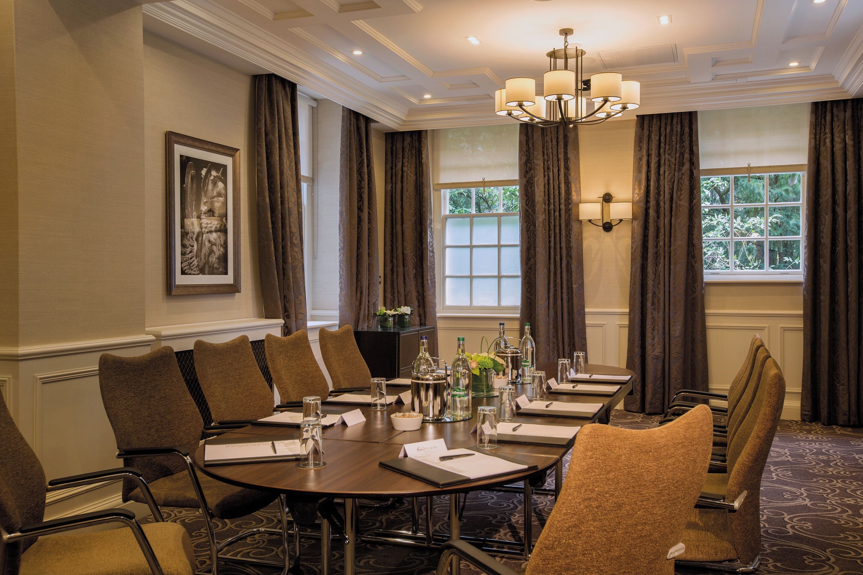 A conference room with a round table, chairs, a chandelier, and a window with curtains.
