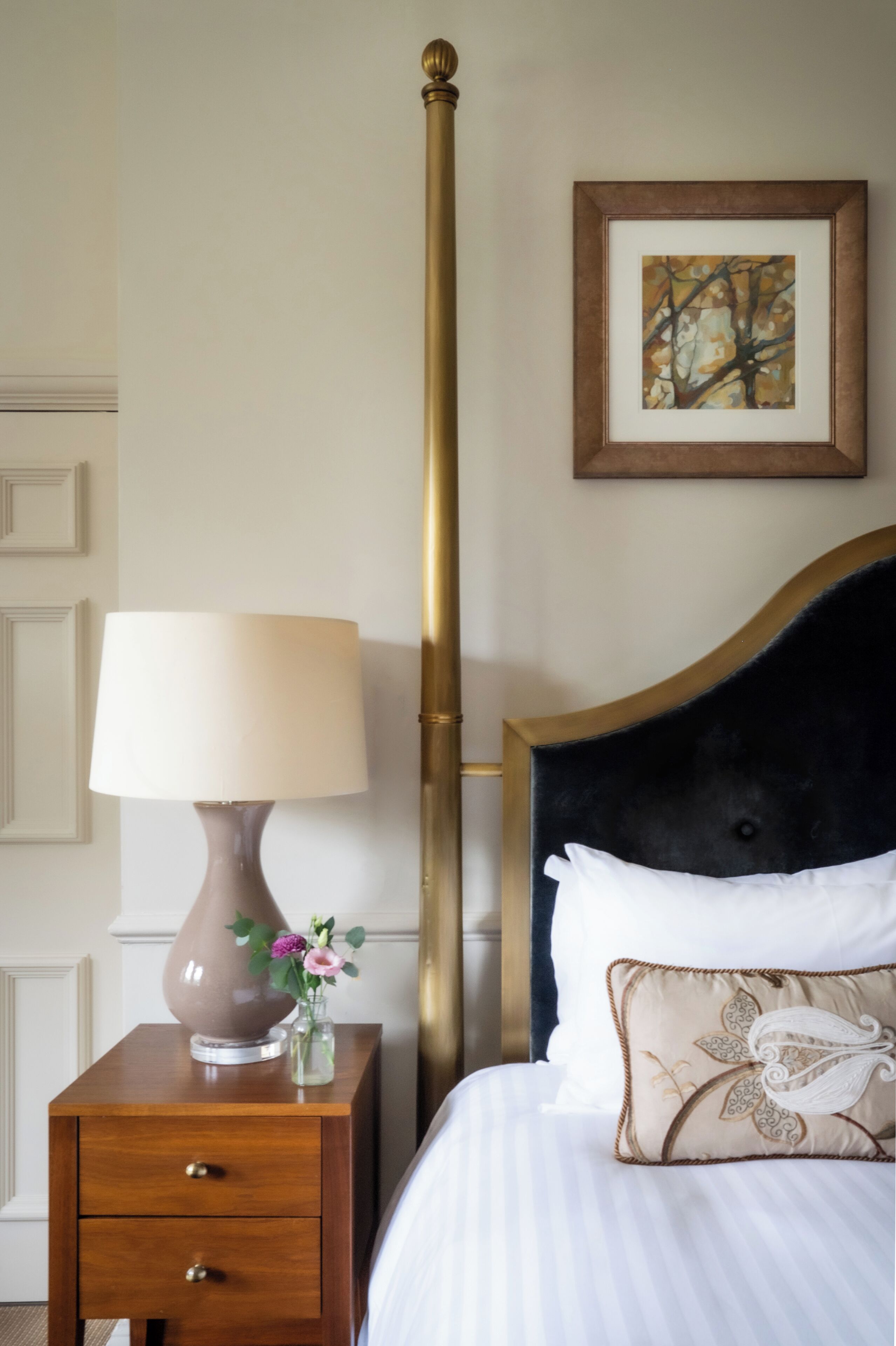A neatly made bed with a headboard, a bedside lamp, a small vase with flowers, and a framed picture on the wall.