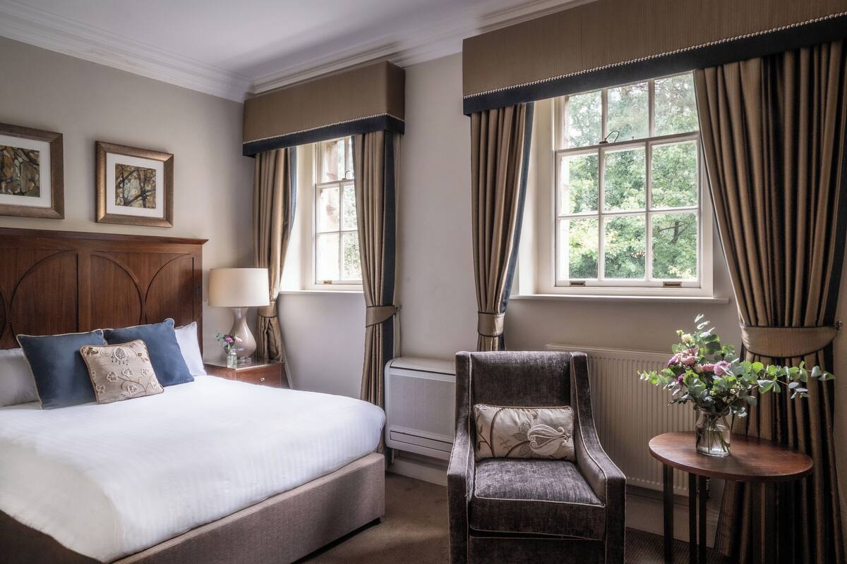 Quarto de hotel bem arrumado com uma cama grande, uma cadeira, uma mesa com um vaso de flores e uma janela com cortinas.