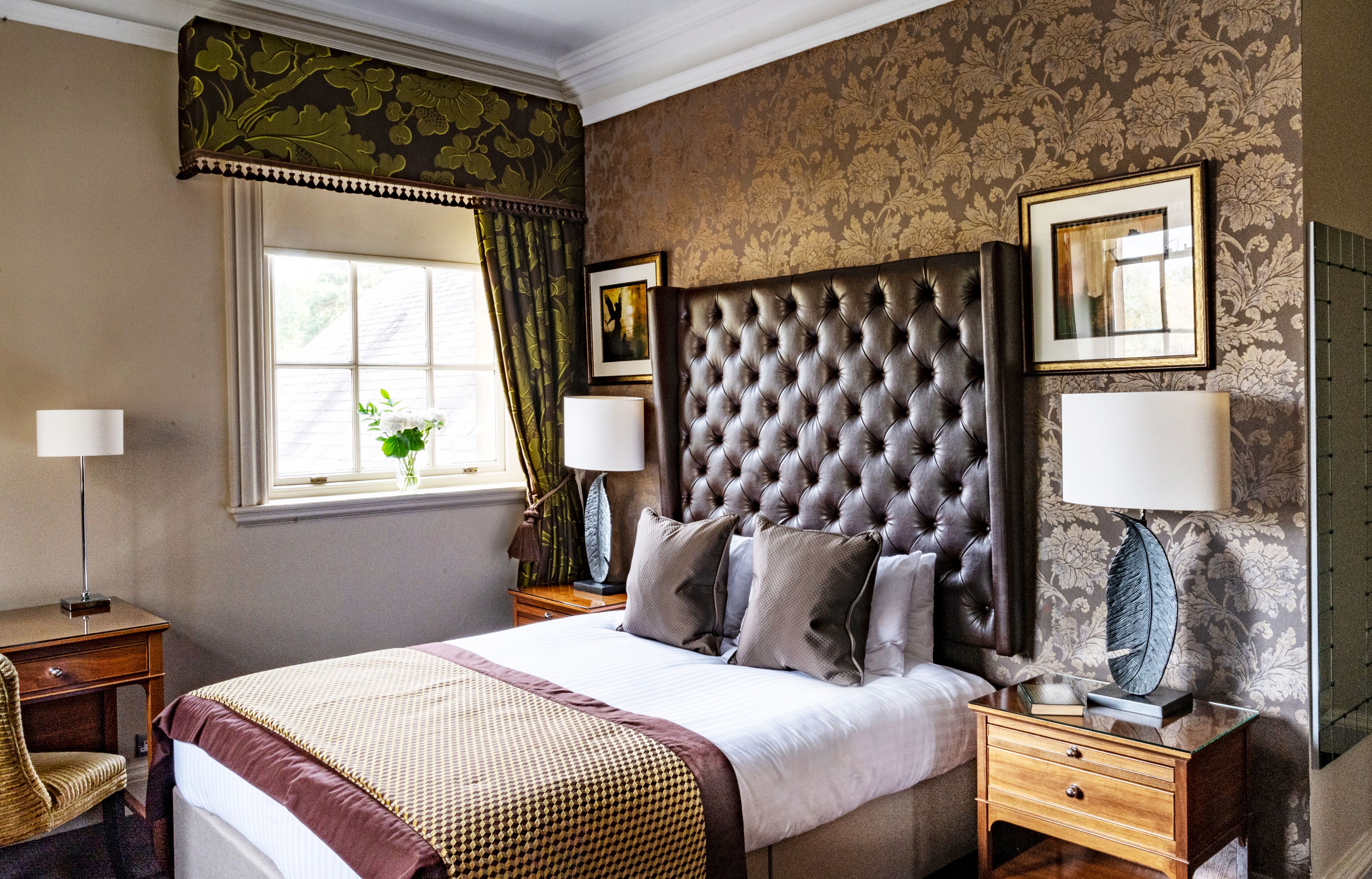 A bedroom with a large bed, two bedside tables, a nightstand, a window with curtains, and a floral wallpaper pattern.