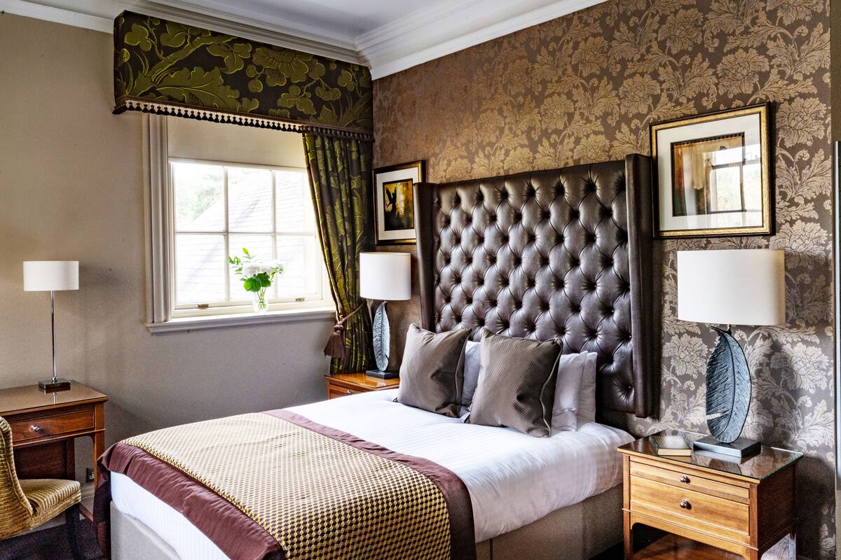 Quarto com uma cama grande, duas mesinhas de cabeceira, um criado-mudo, uma janela com cortinas e um papel de parede estampado com flores.