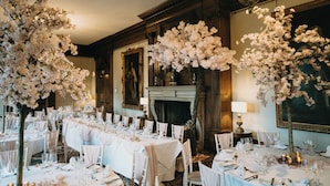 A formal dining room with long tables set for a meal, surrounded by white chairs, and decorated with large white floral arrangements. - Crathorne Hall Hotel (Yarm)