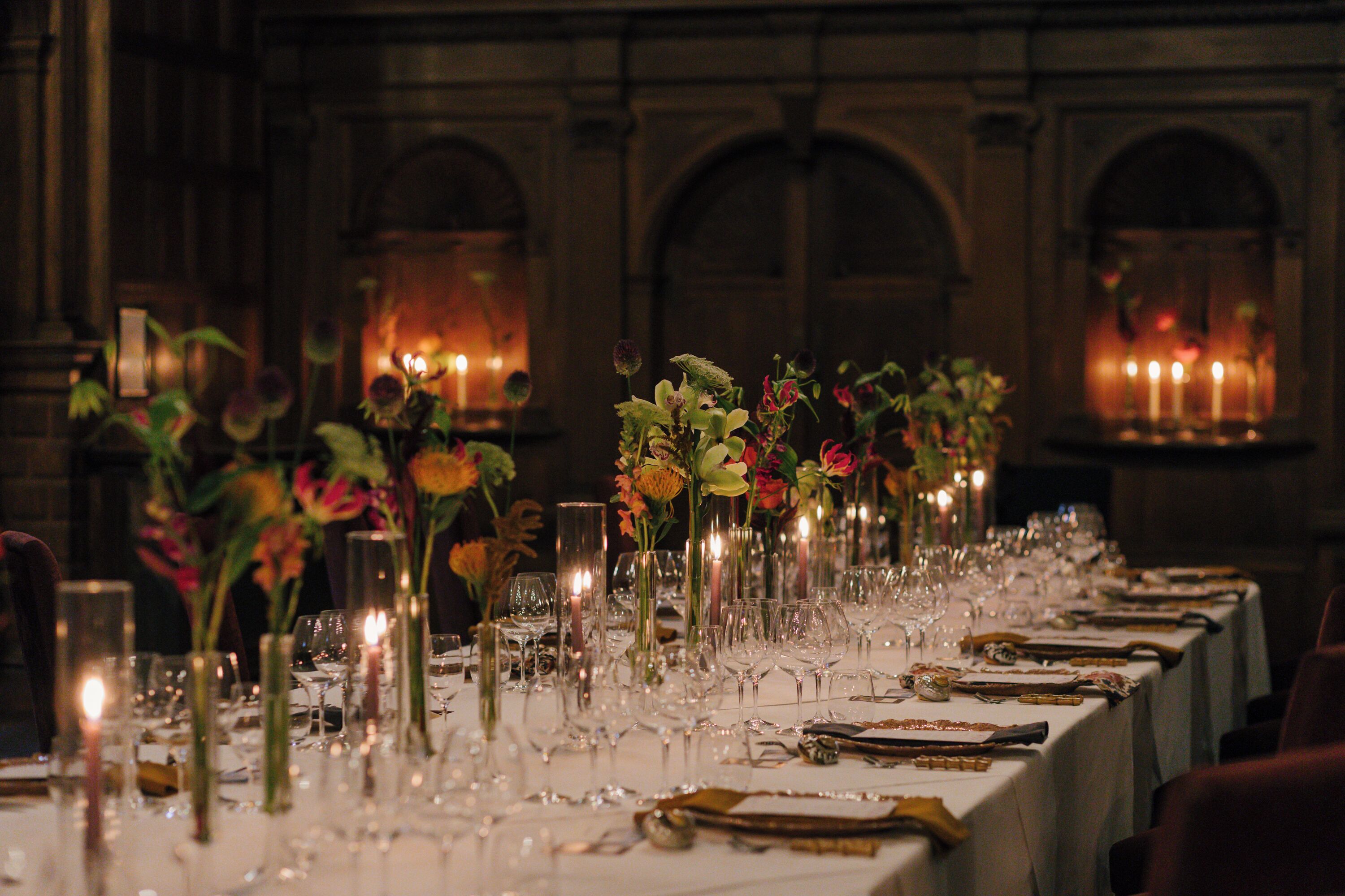 A long dining table set with candles, glassware, and floral centerpieces in a dimly lit room with arched windows.
