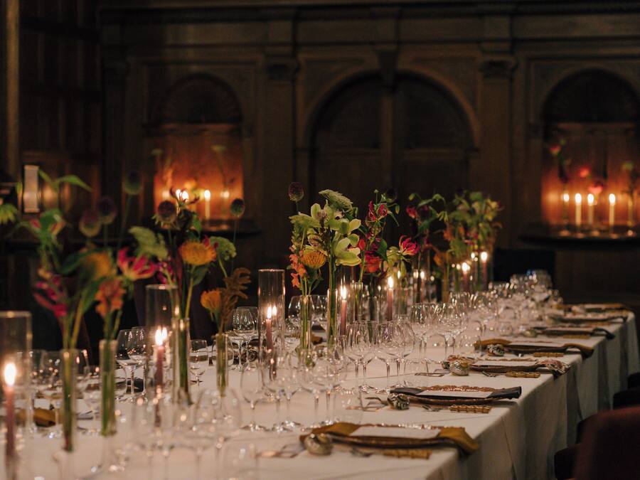 A long dining table set with candles, glassware, and floral centerpieces in a dimly lit room with arched windows.
