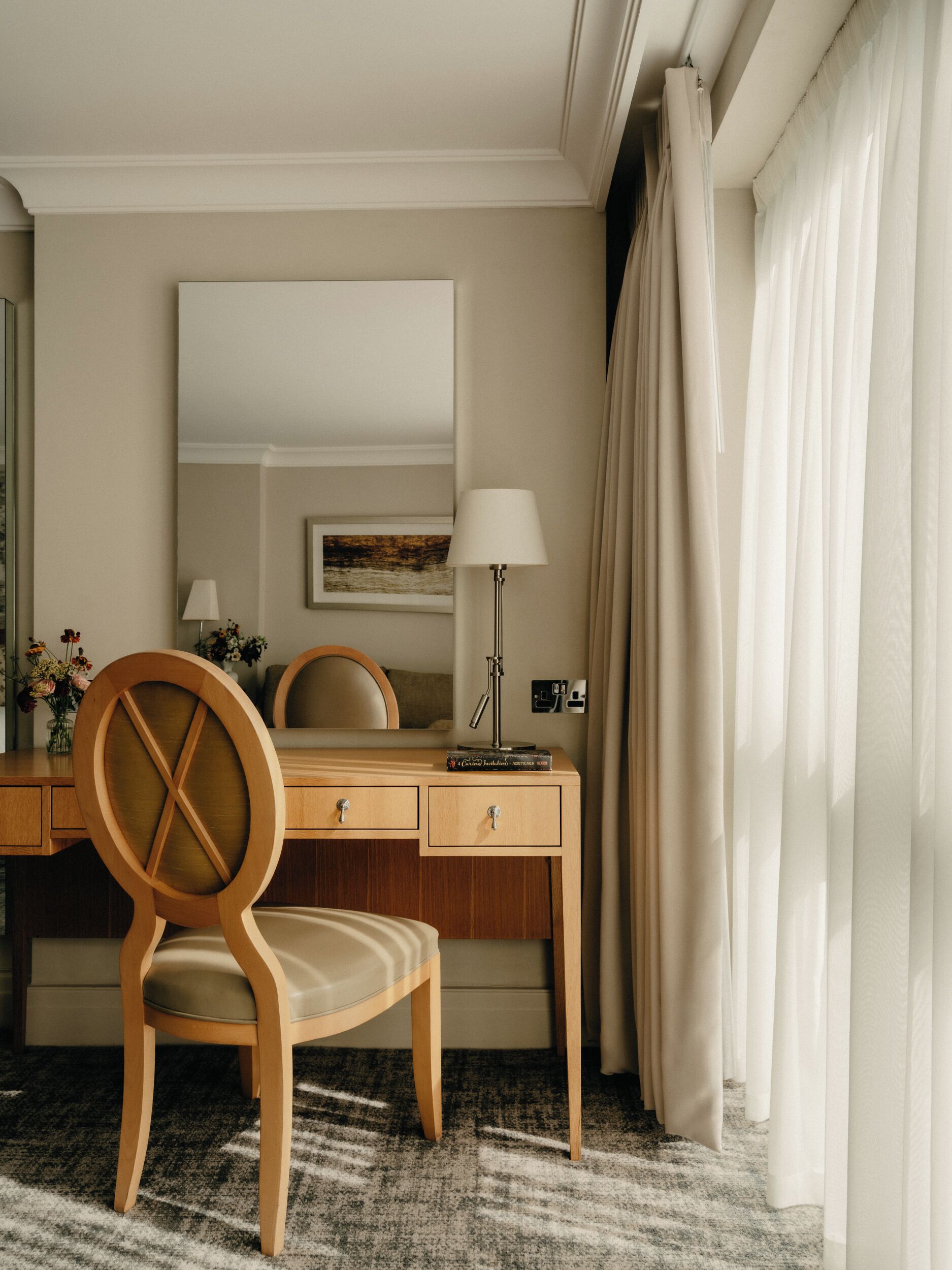Habitación de hotel con un escritorio de madera, una silla, una lámpara, un espejo y una ventana con cortinas.