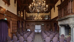 A conference room with rows of chairs, a large chandelier, and a stage with a white screen. - Rhinefield House Hotel (Brockenhurst)