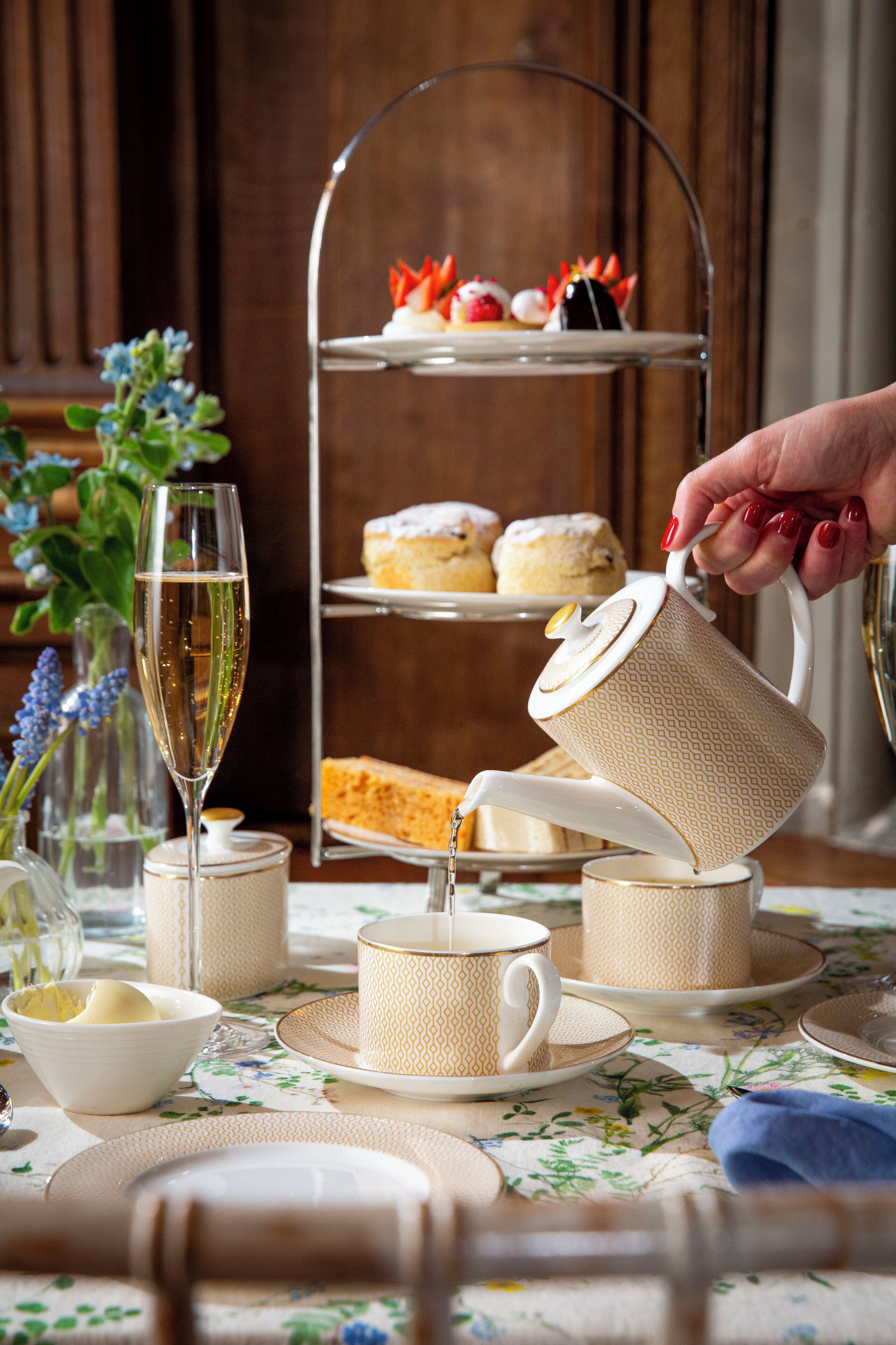 A tiered tea service with a hand pouring tea into a cup, surrounded by floral tablecloth and a glass of champagne.