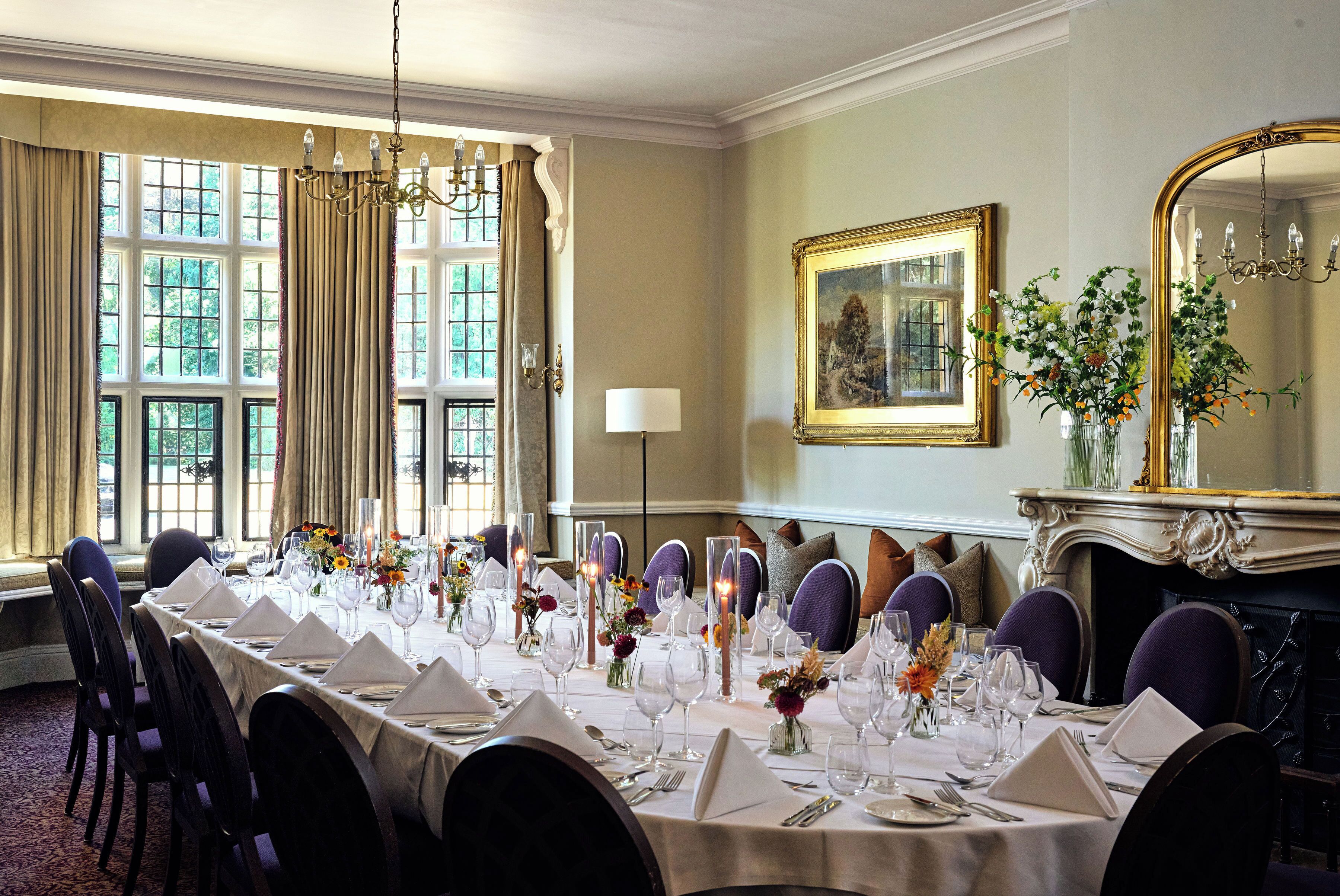 A formal dining room with a long table set for a meal, large windows with curtains, a chandelier, and a fireplace with a mirror.