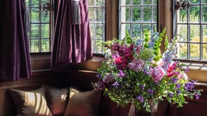 A room with a large window, a wooden bench, and a table with a vase of flowers. - Rhinefield House Hotel (Brockenhurst)