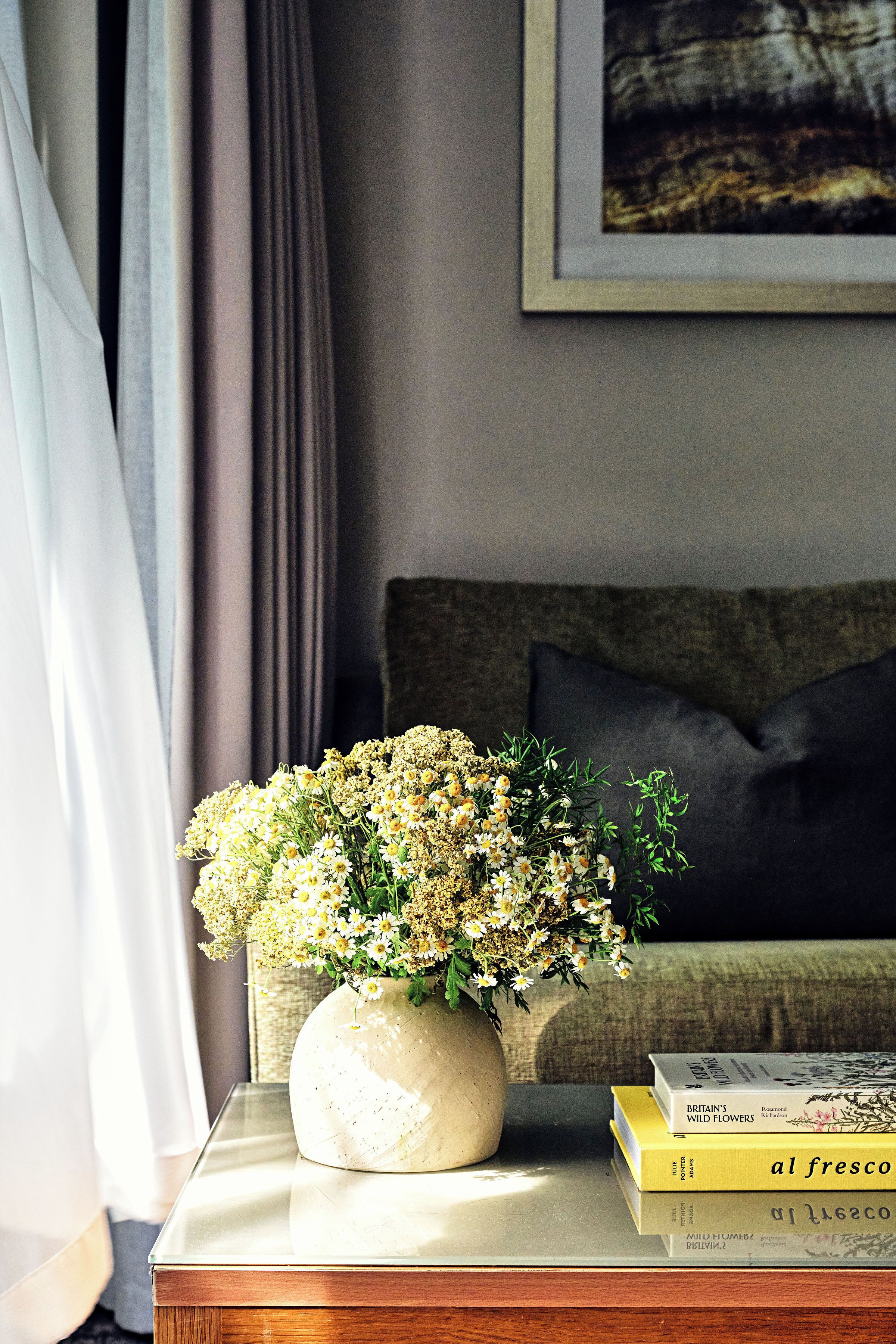 A vase with flowers on a bedside table, a book, and a framed picture on the wall.
