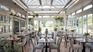 A conservatory with tables and chairs, framed pictures on the walls, and hanging plants.