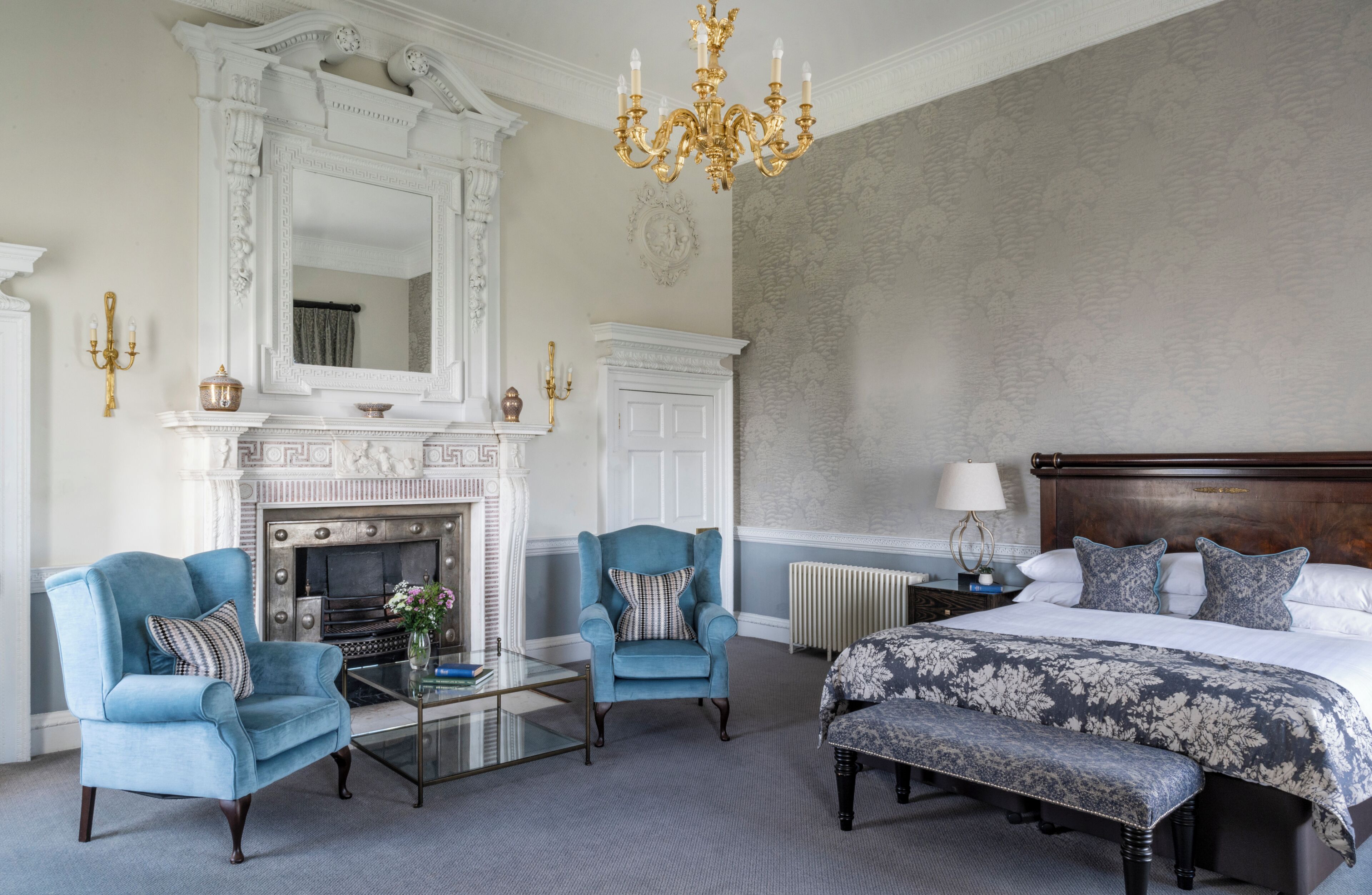 A bedroom with a large bed, two armchairs, a fireplace, and a chandelier.