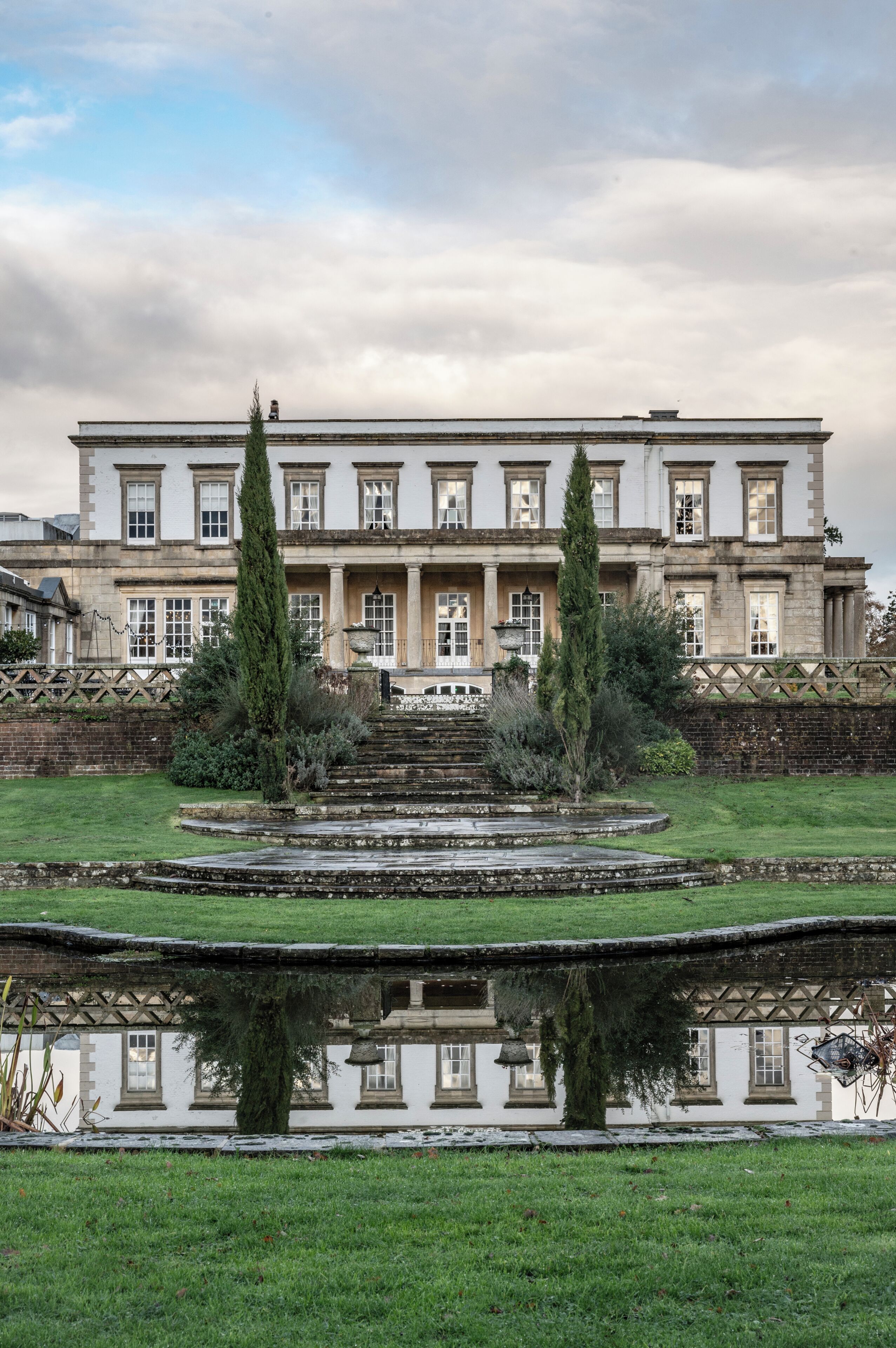 Un edificio grande e histórico con fachada simétrica, entrada central y un jardín circundante con un estanque reflectante.