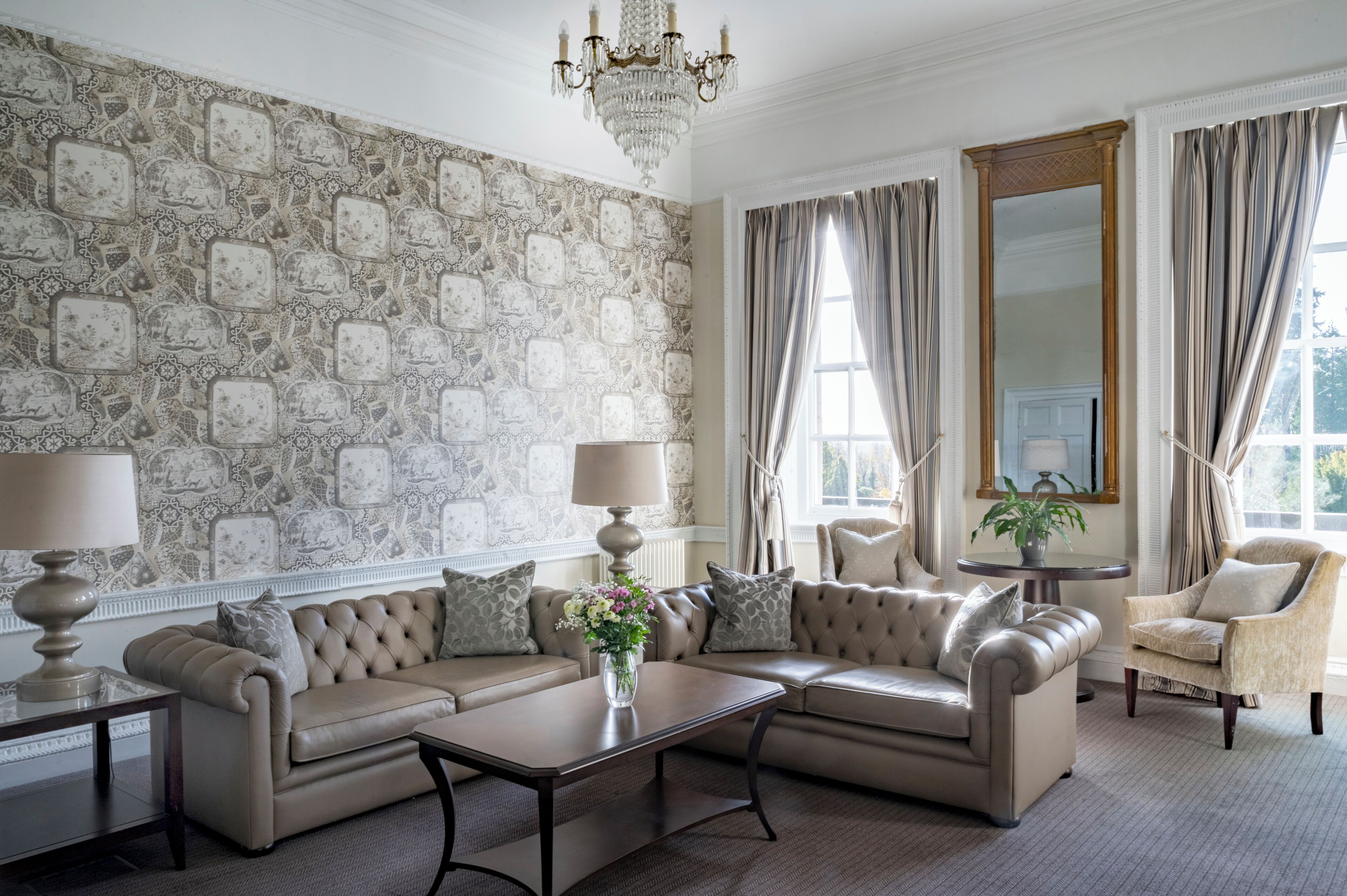 Una habitación con papel tapiz estampado, un candelabro, un conjunto de sofás, una mesa con un jarrón de flores y una silla.