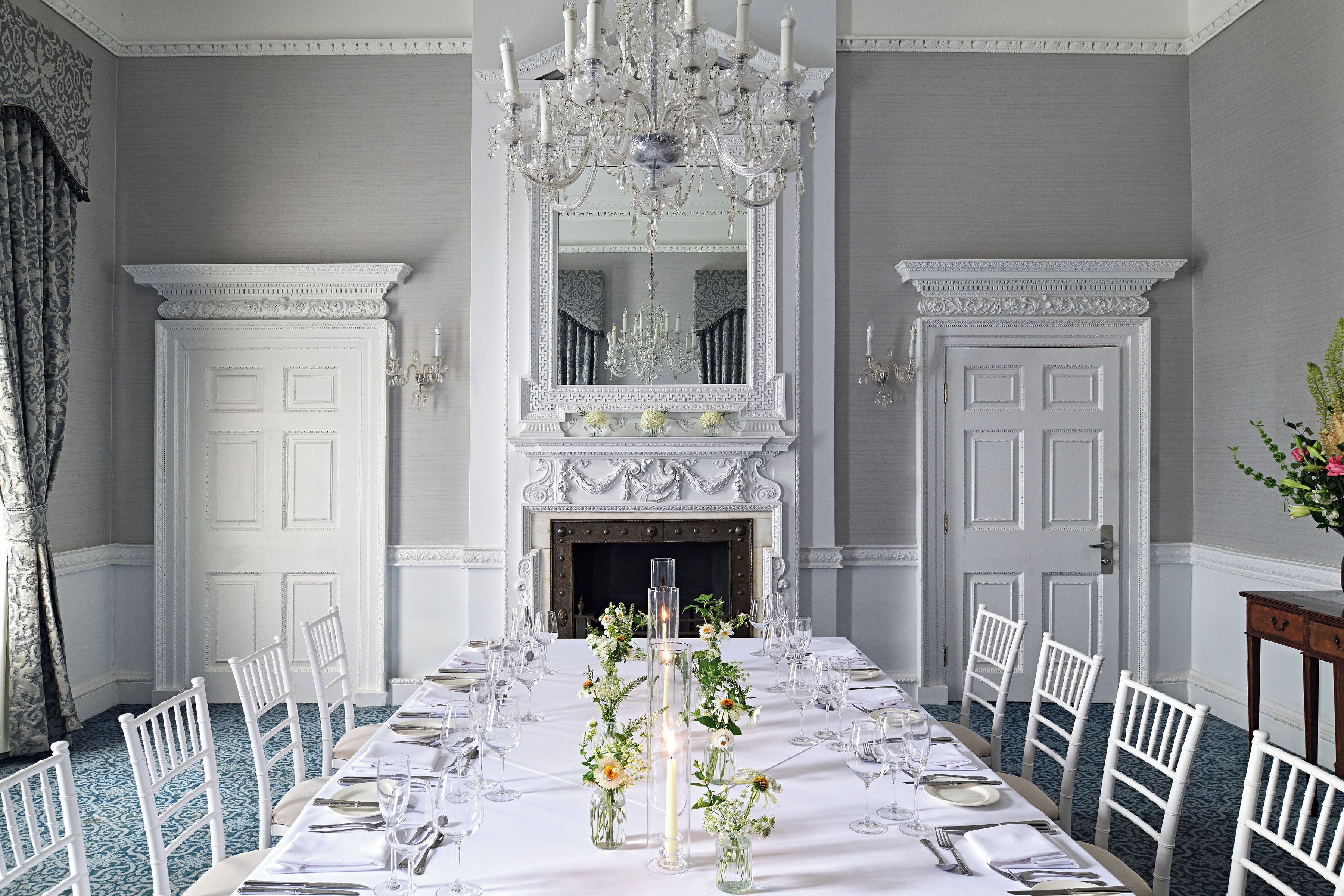 Ein eleganter Speisesaal mit einem langen Tisch, der für eine Mahlzeit gedeckt ist, umrahmt von weißen Stühlen, einem großen Spiegel und einer prunkvollen Chandelier.