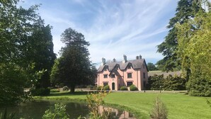 A large, pink building with a pond in the foreground, surrounded by lush greenery. - Cwrt Bleddyn Hotel & Spa (Usk)