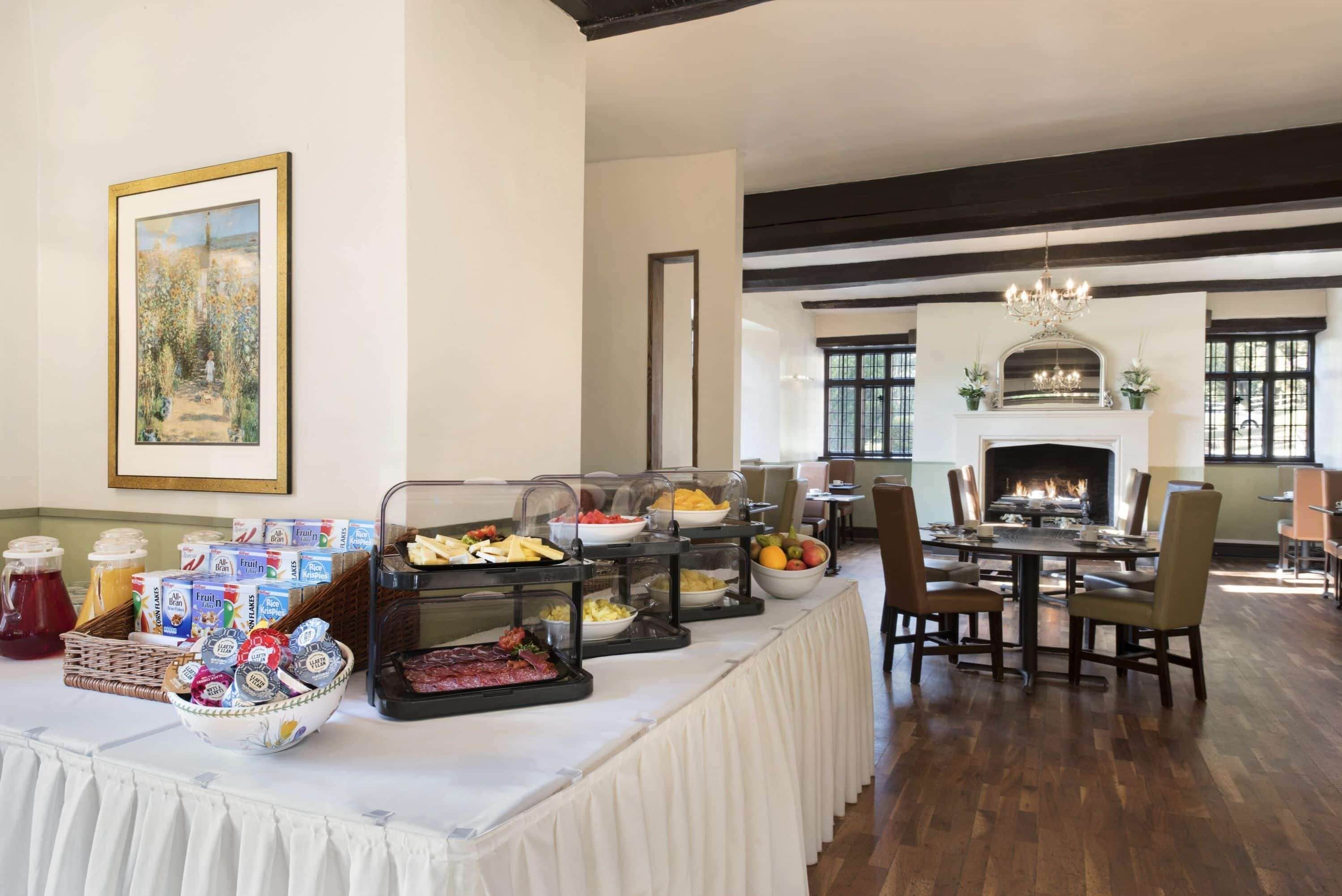 Buffet de café da manhã com diversos alimentos e bebidas, lareira e área de refeições com cadeiras e mesa.