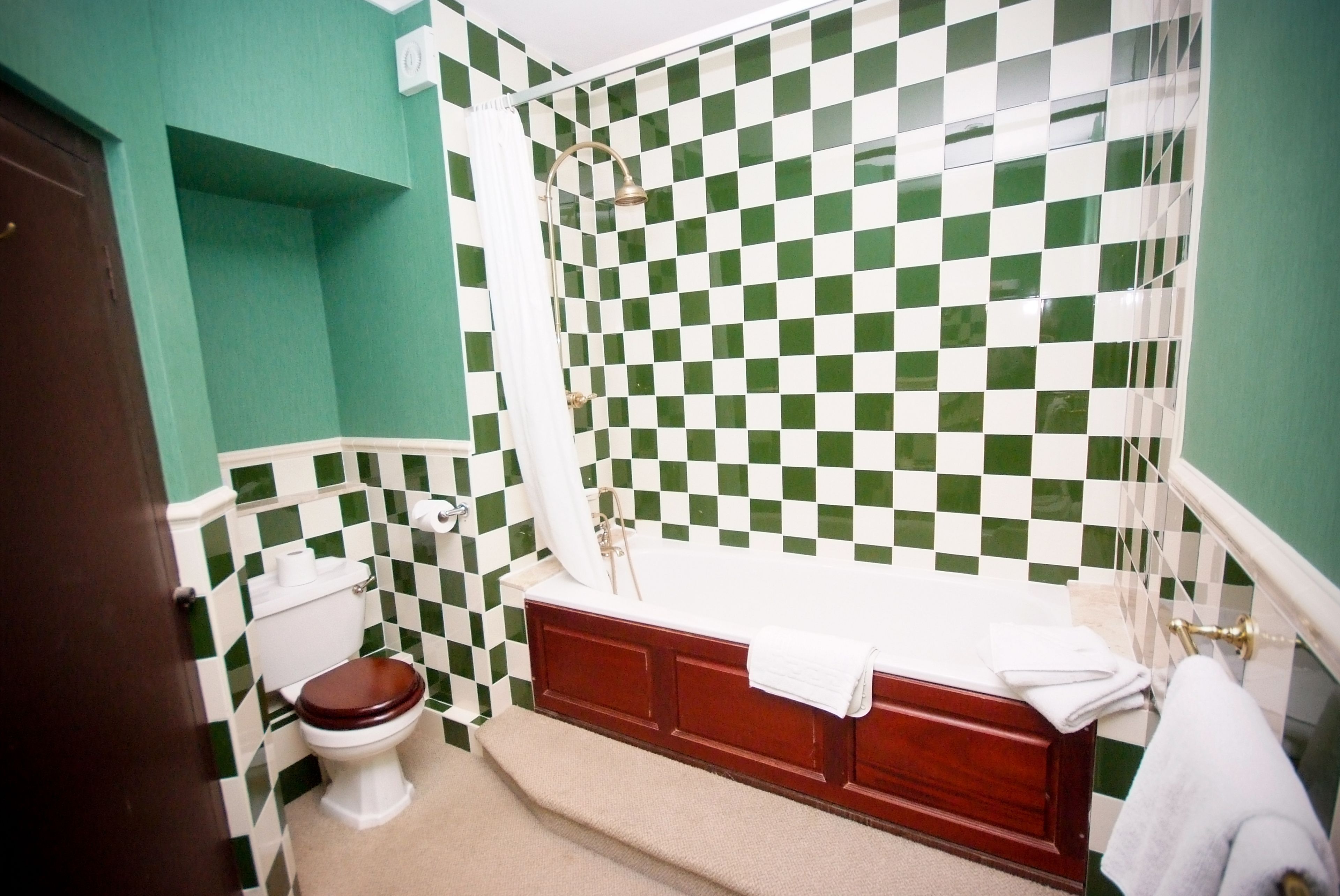 Un baño con pared de azulejos blancos y verdes a cuadros, inodoro blanco, tocador de madera con lavamanos blanco y bañera con cortina de ducha.