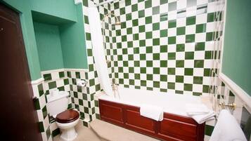 Un baño con pared de azulejos blancos y verdes a cuadros, inodoro blanco, tocador de madera con lavamanos blanco y bañera con cortina de ducha.