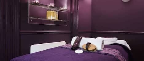 A spa room with a purple color scheme, a bed with a white headboard, a mirror with shelves, and a wall-mounted light labeled "ESPA".
