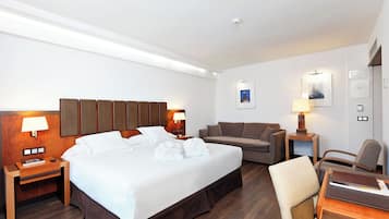 A hotel room with a large bed, a sofa, a desk, and a chair. There are two bedside tables with lamps and a wall-mounted shelf.