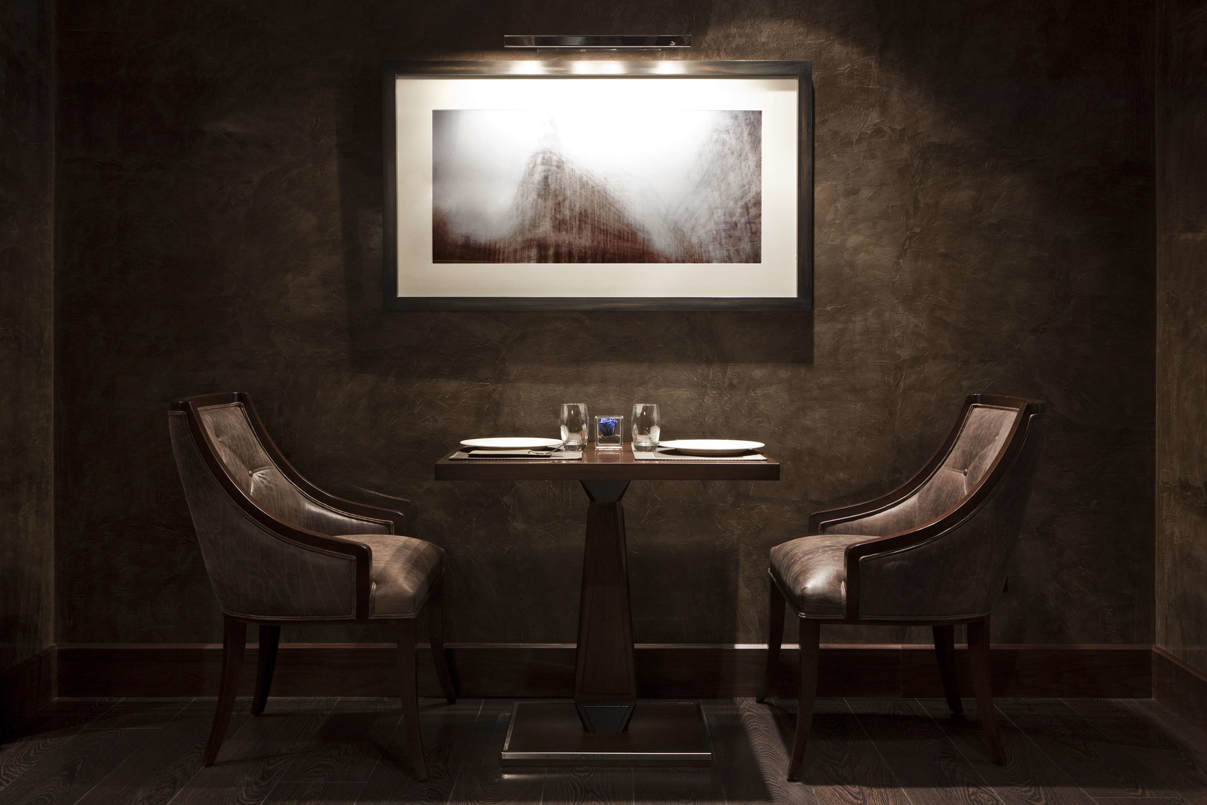 Comedor de ambiente oscuro con una mesa y dos sillones de cuero, una obra enmarcada sobre la mesa y una pared texturizada oscura.