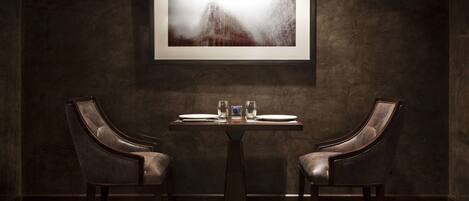 A dark-themed dining area with a table and two leather chairs, a framed artwork above the table, and a dark textured wall.