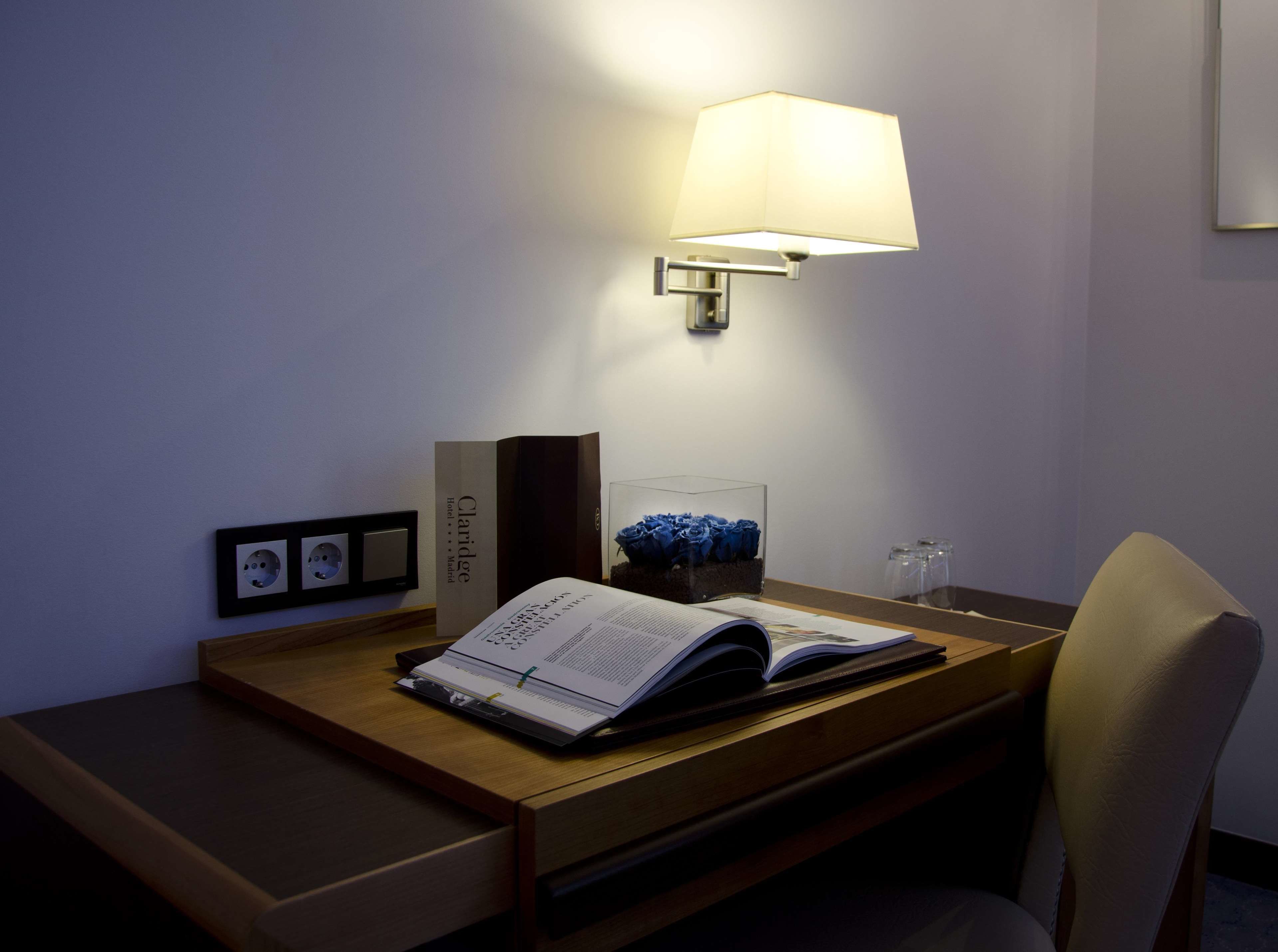 Een bureau in een hotelkamer met een lamp, een boek, een glazen vaas met blauwe rozen en een lijstje aan de muur.