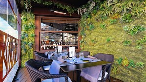 Un área de comedor al aire libre con sillas y mesas de mimbre, una terraza de madera y una pared cubierta de vegetación.