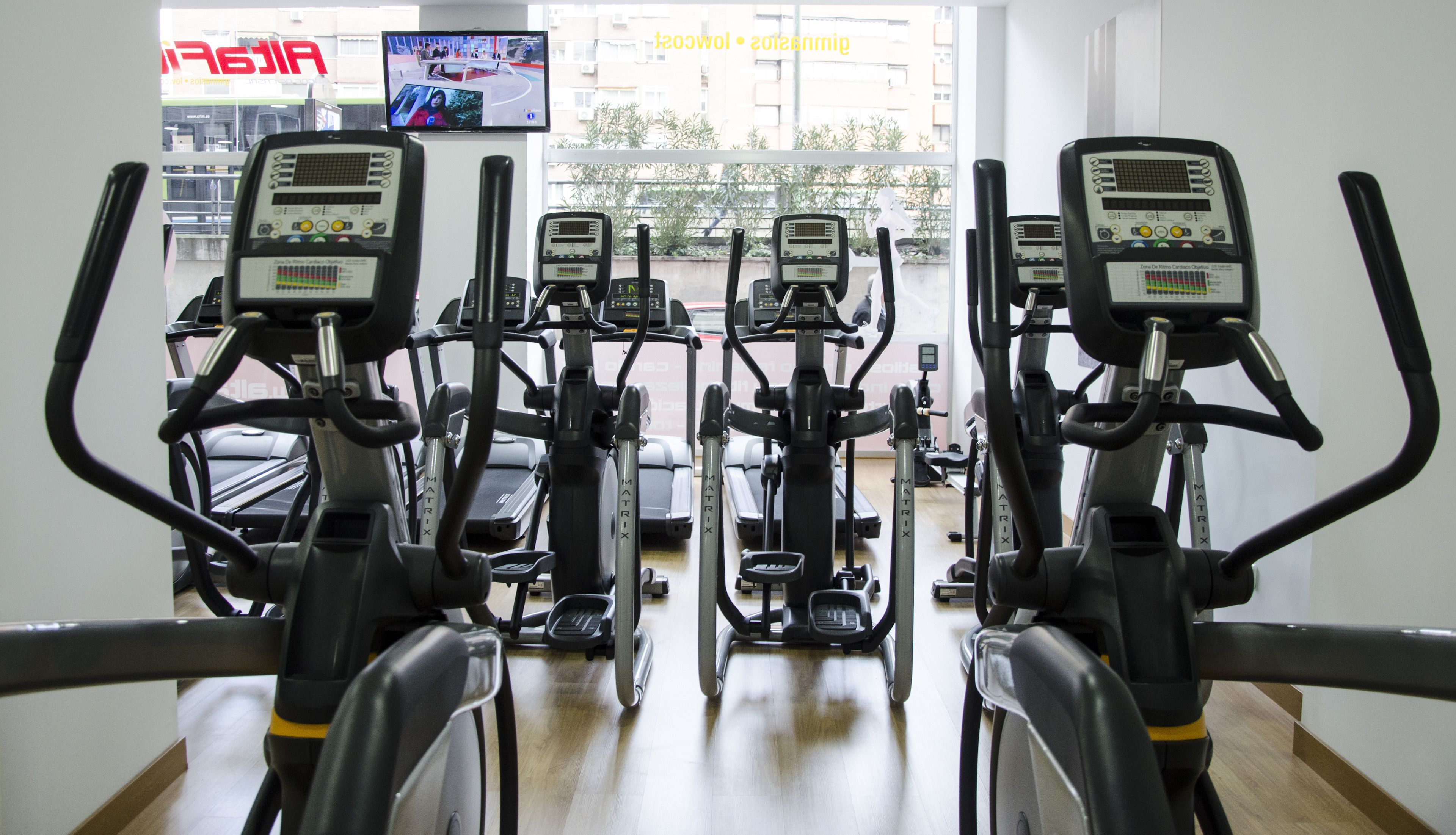 A gym with multiple stationary bikes and treadmills, a TV screen displaying a video, and a view of the outside through large windows.