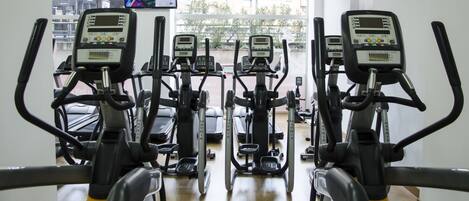 A gym with multiple stationary bikes and treadmills, a TV screen displaying a video, and a view of the outside through large windows.