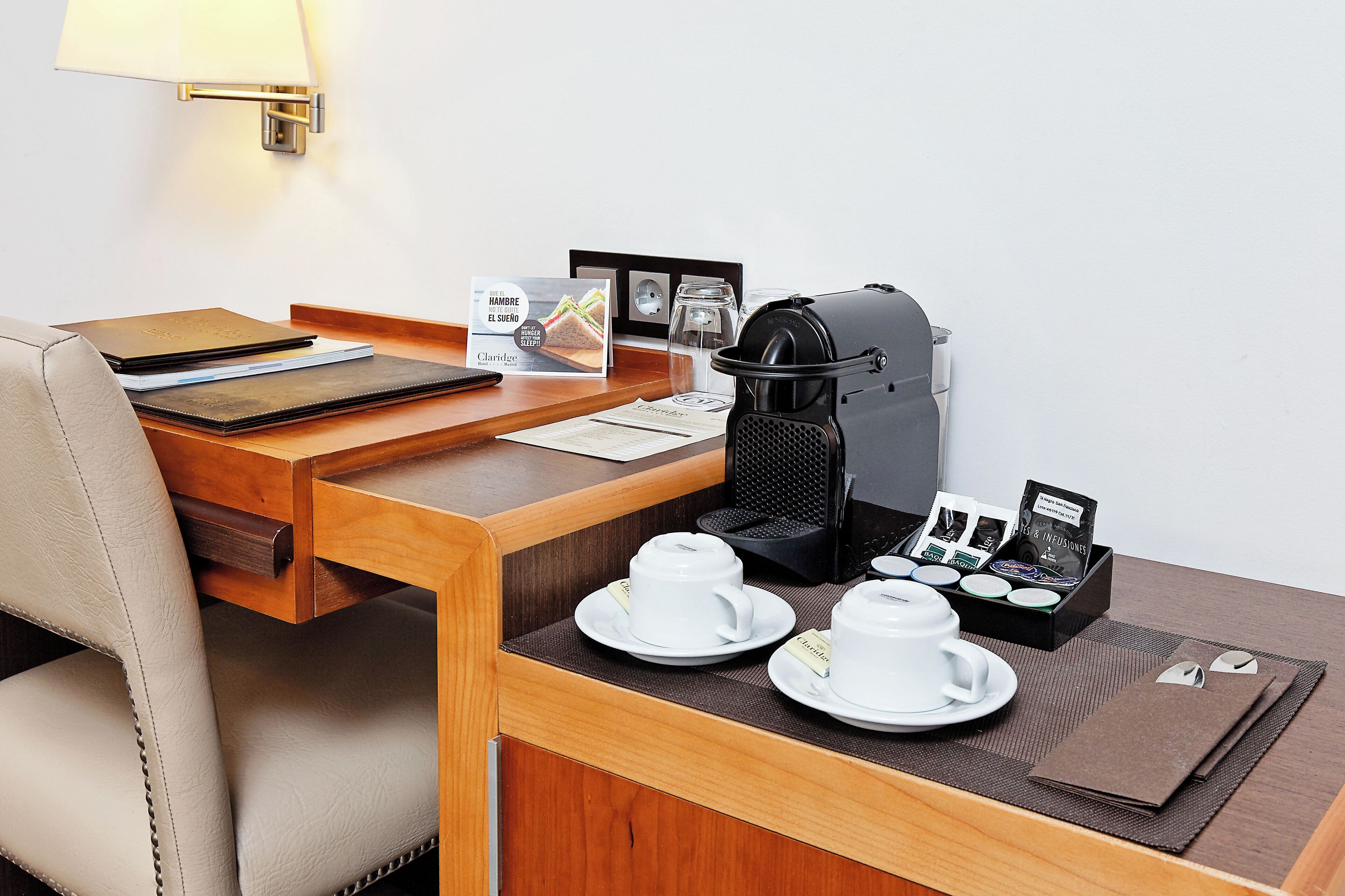 Habitació d'hotel amb escriptori de fusta, un ordinador portàtil, una cafetera i un safata de petit esmorzar amb tasses i una teiera.