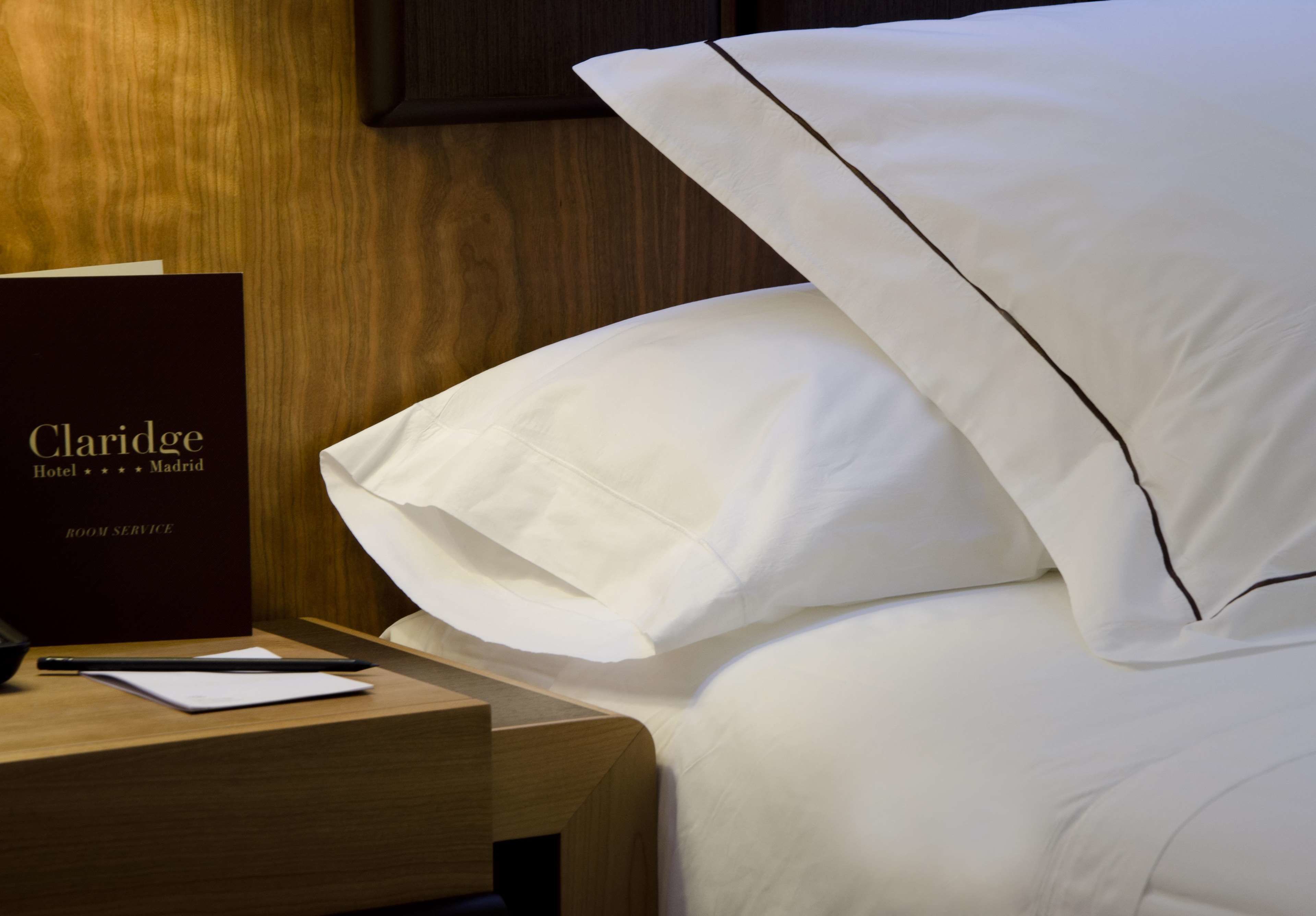 Une chambre d’hôtel avec une tête de lit en bois, du linge de lit blanc et une enseigne indiquant « Claridge Hotel ++ Madrid ».