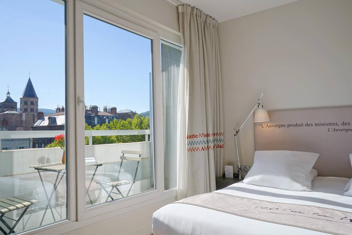 Una habitación de hotel con un ventanal grande, una cama, una mesita de noche y una lámpara.