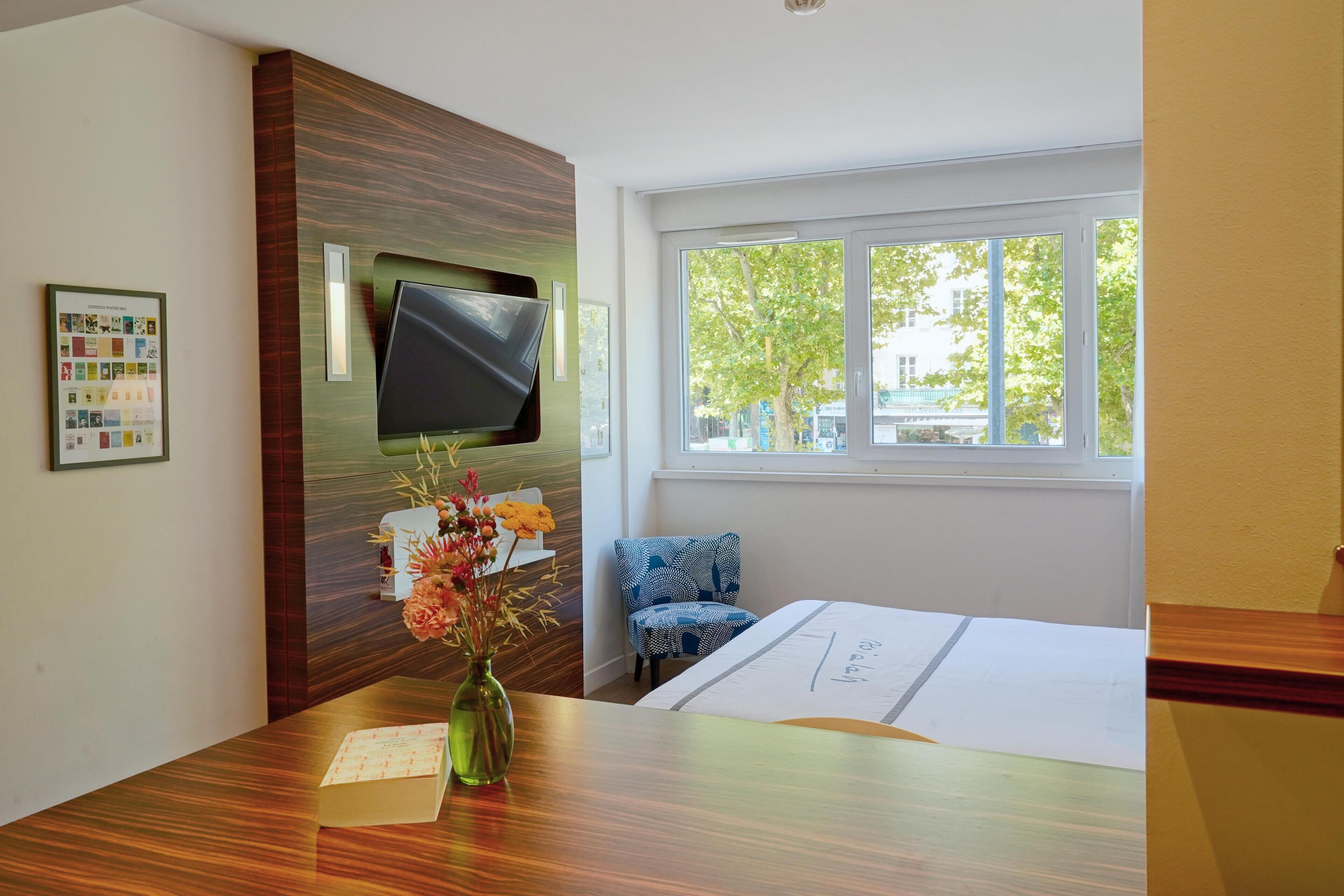 A hotel room with a wooden reception desk, a flat-screen TV mounted on a wooden panel, a window with a view of trees, a blue patterned chair, and a bed with a white bedspread.