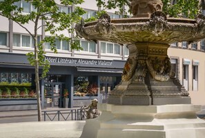 A fountain with intricate carvings, a building with a sign reading "Hôtel Littéraire Alexandre Vialatte," and a tree in the foreground.